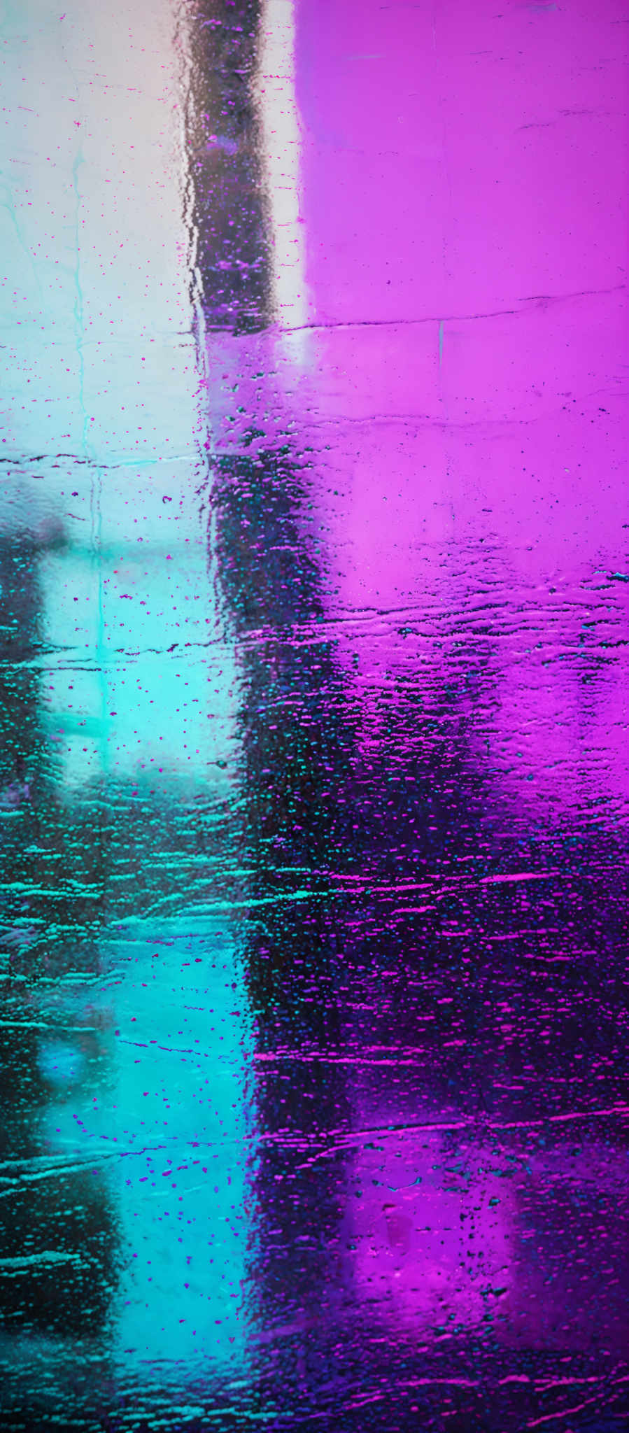 The image showcases a vibrant and colorful reflection on a wet surface. The dominant colors are shades of blue, purple, and pink. The reflection appears to be of a cityscape or urban environment, with tall structures and lights. The surface has a glossy appearance, suggesting it might be a wet street or pavement, possibly after a rain. The reflections are slightly distorted, indicating movement or the presence of wind.