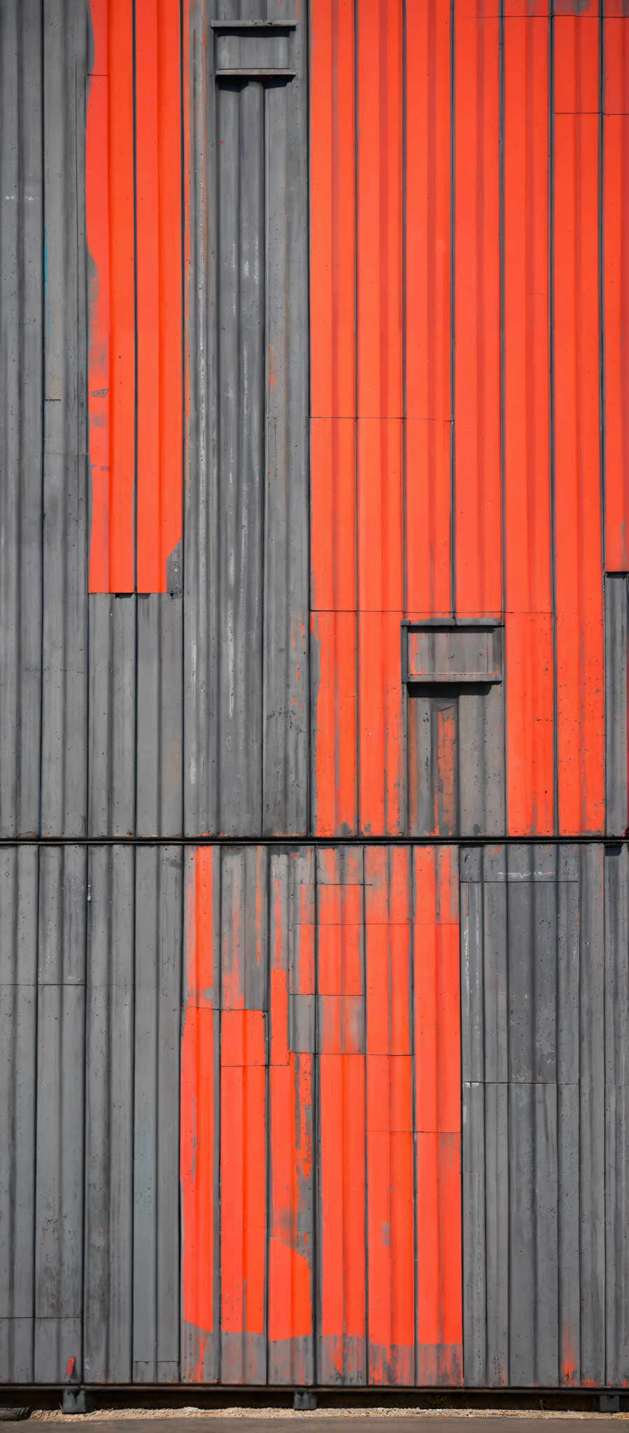 The image showcases a vertical structure with alternating sections of gray and bright orange. The gray sections appear weathered, with visible wear and tear, including chipped paint and rust. The orange sections are more vibrant and seem to be newer or less exposed to external elements. The structure has vertical lines, and there are rectangular cutouts or openings in the gray sections.