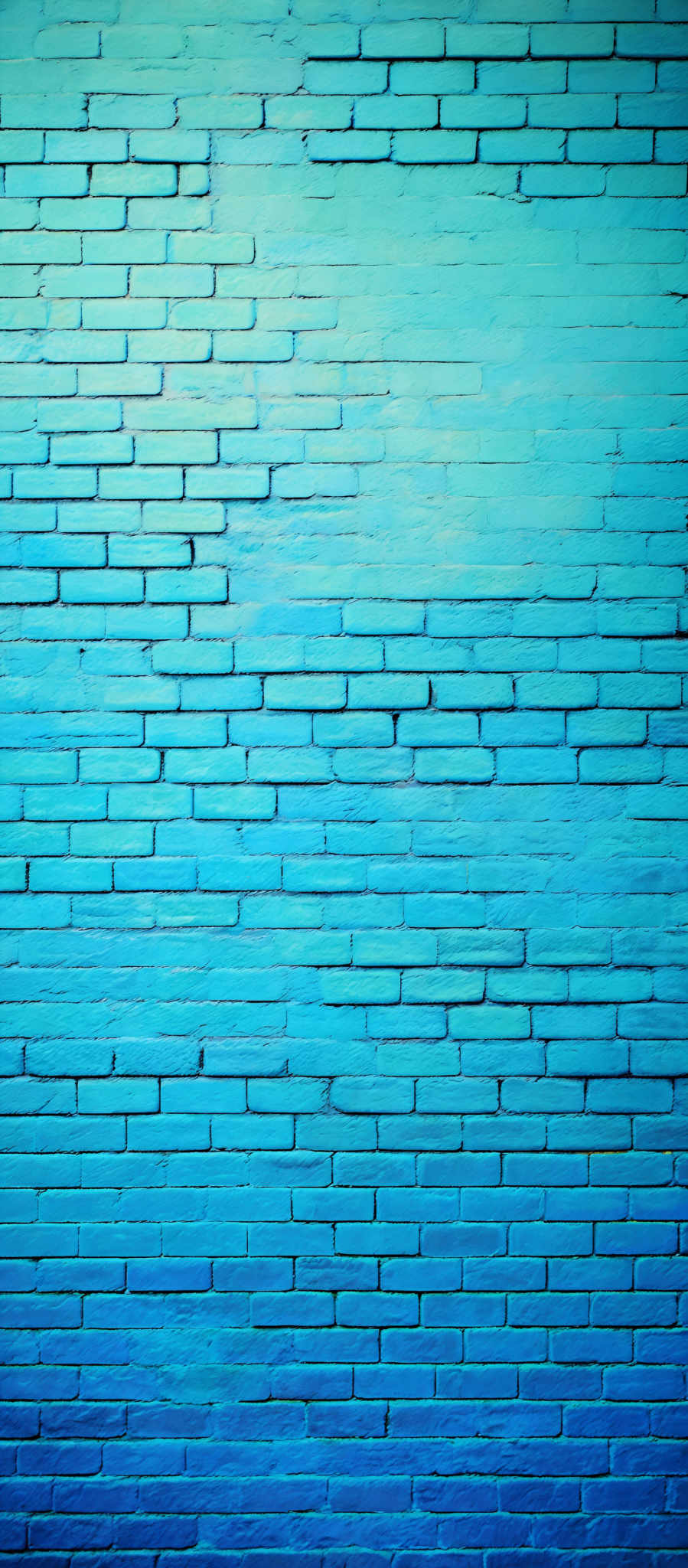 The image showcases a brick wall painted in varying shades of blue. The bricks are rectangular in shape and are arranged in a staggered pattern. The color gradient starts from a light blue at the top and transitions to a deeper shade of blue towards the bottom. The texture of the bricks is visible, and there are slight variations in the shading, giving depth and dimension to the wall.