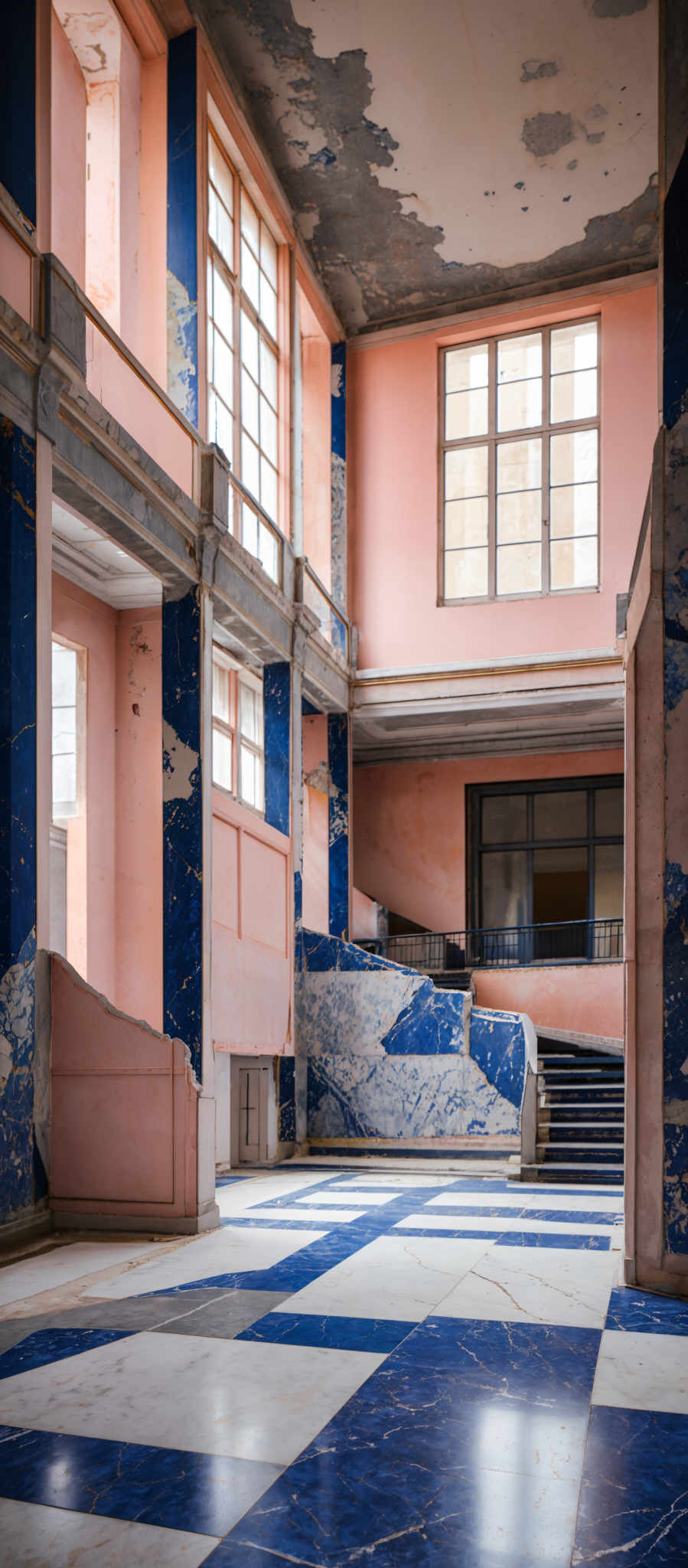 The image showcases an interior space with a combination of pink and blue walls. The ceiling has a peeling paint, revealing patches of white and gray. The floor features a checkered pattern with alternating white and blue tiles. Large windows allow natural light to flood the space, illuminating the peeling walls and the checkered floor. The architectural design includes columns and staircases, with the stairs leading to an upper level.