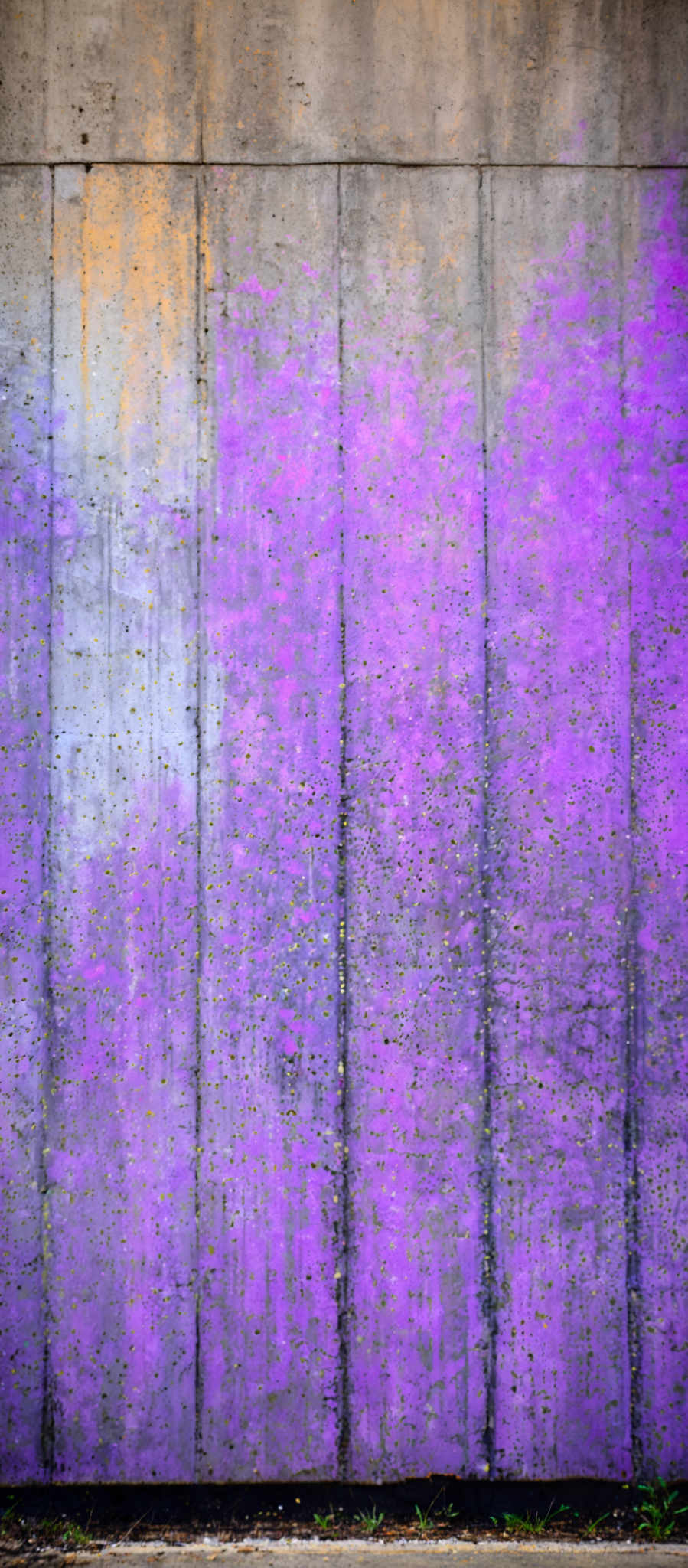 The image showcases a large, textured wall made of what appears to be concrete. The wall has a gradient of colors, transitioning from a light gray at the top to a vibrant purple in the middle. The purple section has a rough texture with numerous small holes and irregularities. At the bottom, there's a section of the wall that is more worn out, showing signs of wear and tear, with patches of discoloration and some moss or lichen growth.