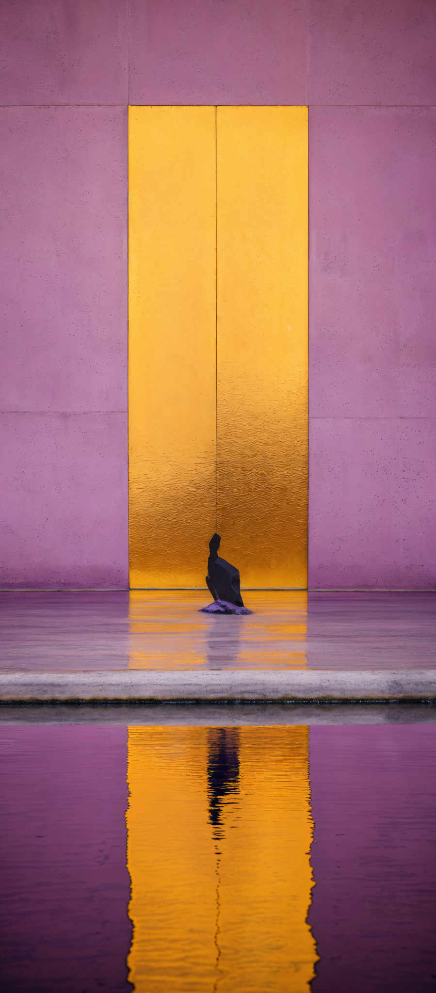 The image showcases a large rectangular golden door set against a pink wall. The door appears to be made of a reflective material, as it mirrors the pink walls and the sky above. In front of the door, there's a calm body of water that reflects the door and the surrounding colors. On the water's surface, there is a silhouette of a person, possibly sitting or kneeling, with a purple object beside them. The overall color palette is dominated by shades of pink, gold, and purple, creating a serene and contemplative atmosphere.