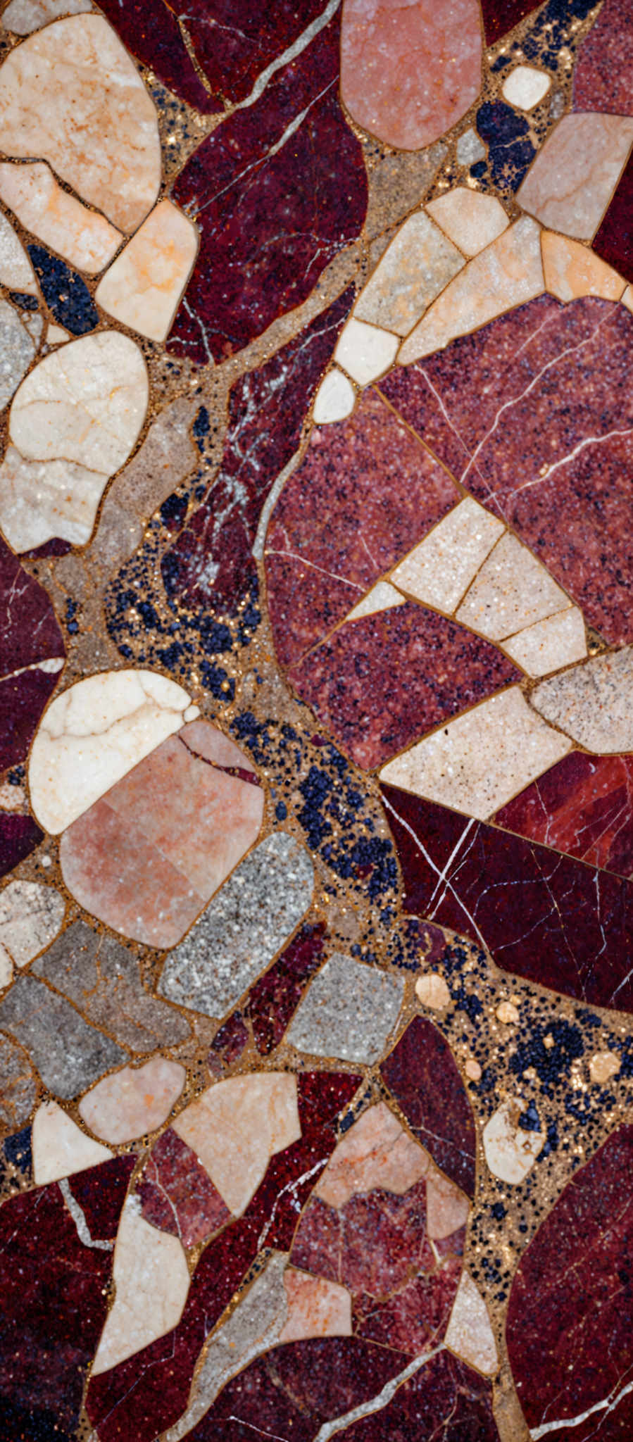 The image showcases a mosaic of various shapes and colors. The predominant colors are shades of red, beige, and gray. The shapes are irregular and resemble fragments of rocks or stones, interspersed with gold-colored streaks. The overall pattern gives an impression of a geological or natural texture, possibly representing a section of a rock or a terrain.