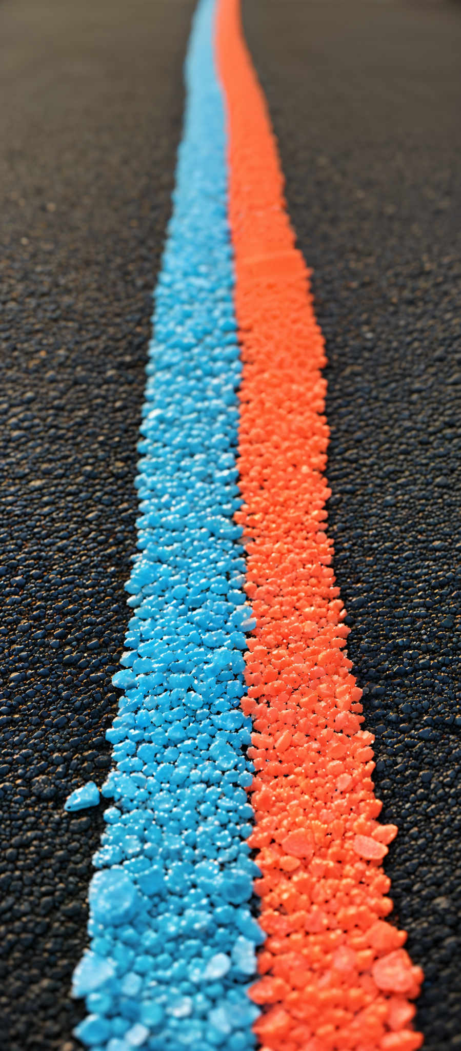 The image showcases a road with a distinct vertical stripe of two colors. The stripe starts from the bottom of the image and extends upwards. The left side of the stripe is painted in a vibrant blue color, while the right side is in a bright orange. The texture of the road appears to be asphalt with a slightly rough surface. The blue and orange stripes are made up of numerous small, rounded gravel or stones, giving them a textured appearance.