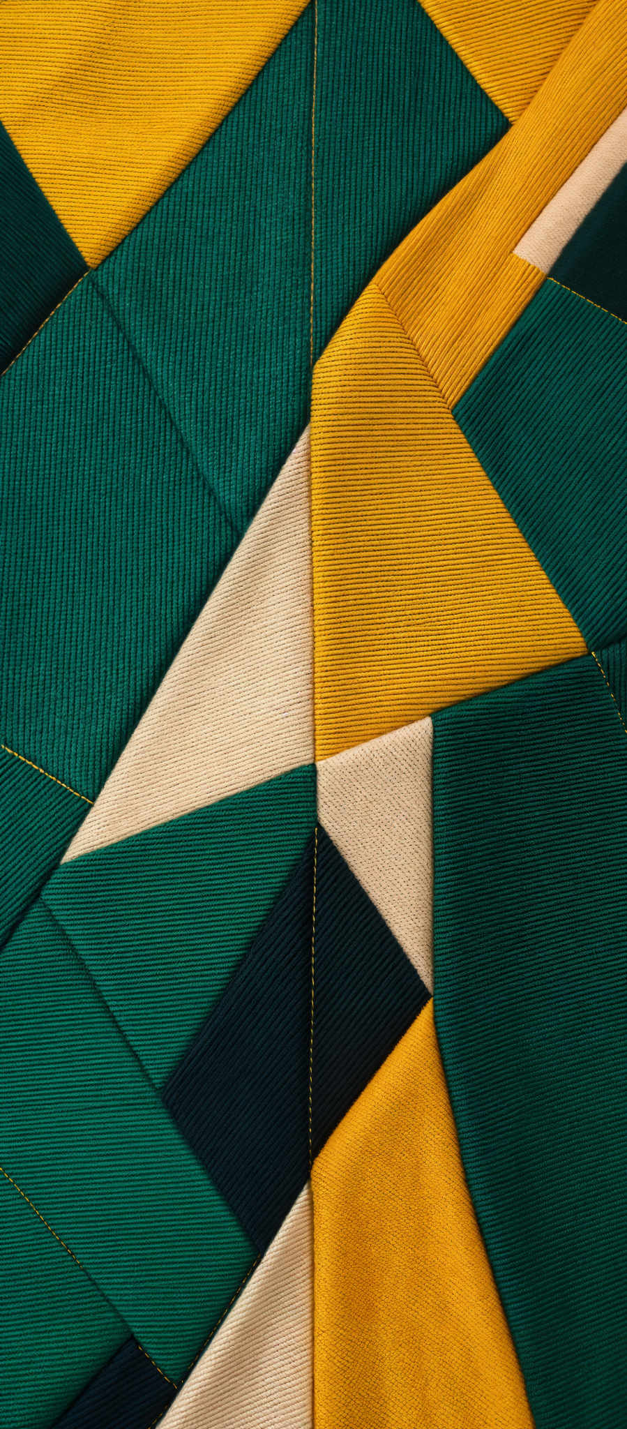 The image showcases a vibrant and intricate pattern made up of geometric shapes. The dominant colors are shades of green, yellow, and beige. The shapes appear to be a combination of triangles and rectangles, sewn together in a patchwork fashion. The texture of the fabric suggests a ribbed or corduroy pattern, and the stitching is meticulous, with visible threads running through the fabric.