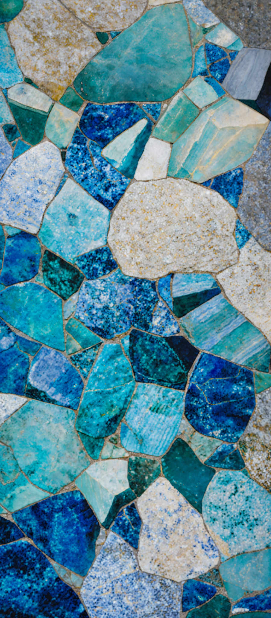 The image showcases a mosaic of irregularly shaped stones or tiles, predominantly in shades of blue, teal, and gray. The stones are interlocked together, forming a pattern that resembles a sea or ocean floor. The varying shades and textures of the stones give the mosaik a natural and organic appearance.