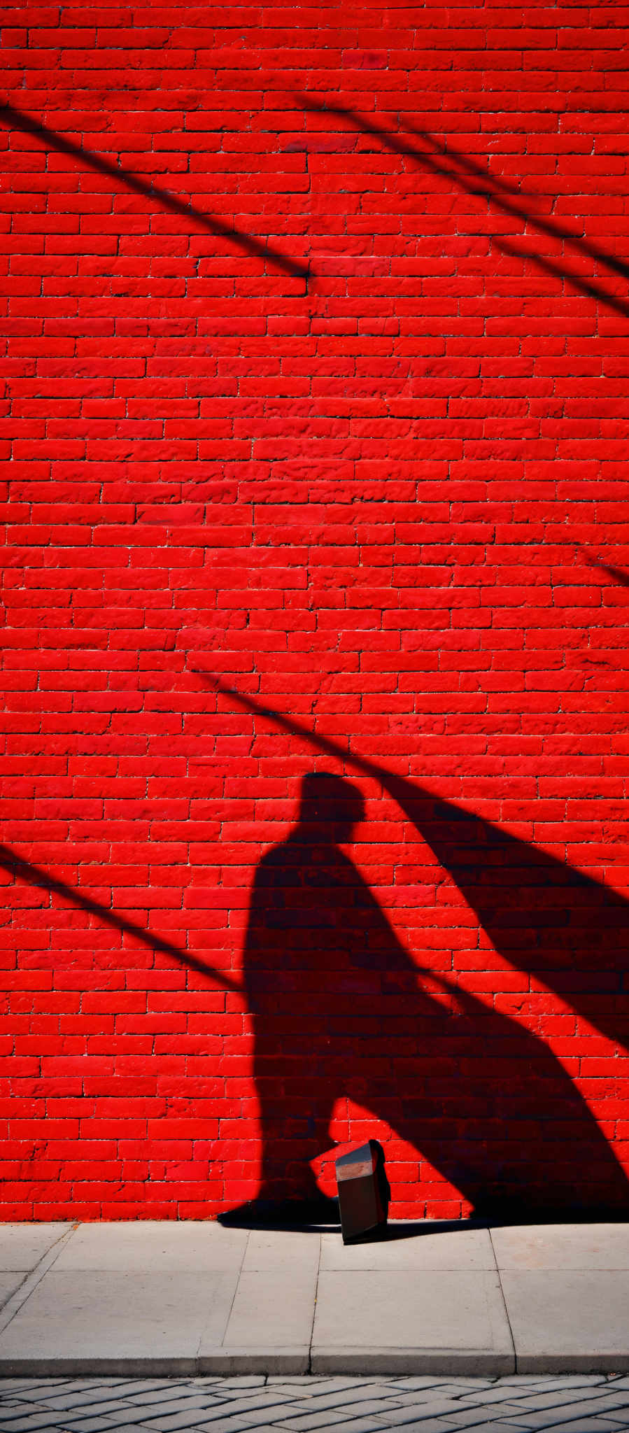 The image showcases a vibrant red brick wall. The sun casts a shadow of a person, possibly walking, on the wall. This shadow is elongated and appears as if the person is moving at a slight angle. There's also a small object, possibly a bag or a briefcase, lying on the ground near the shadow's base.