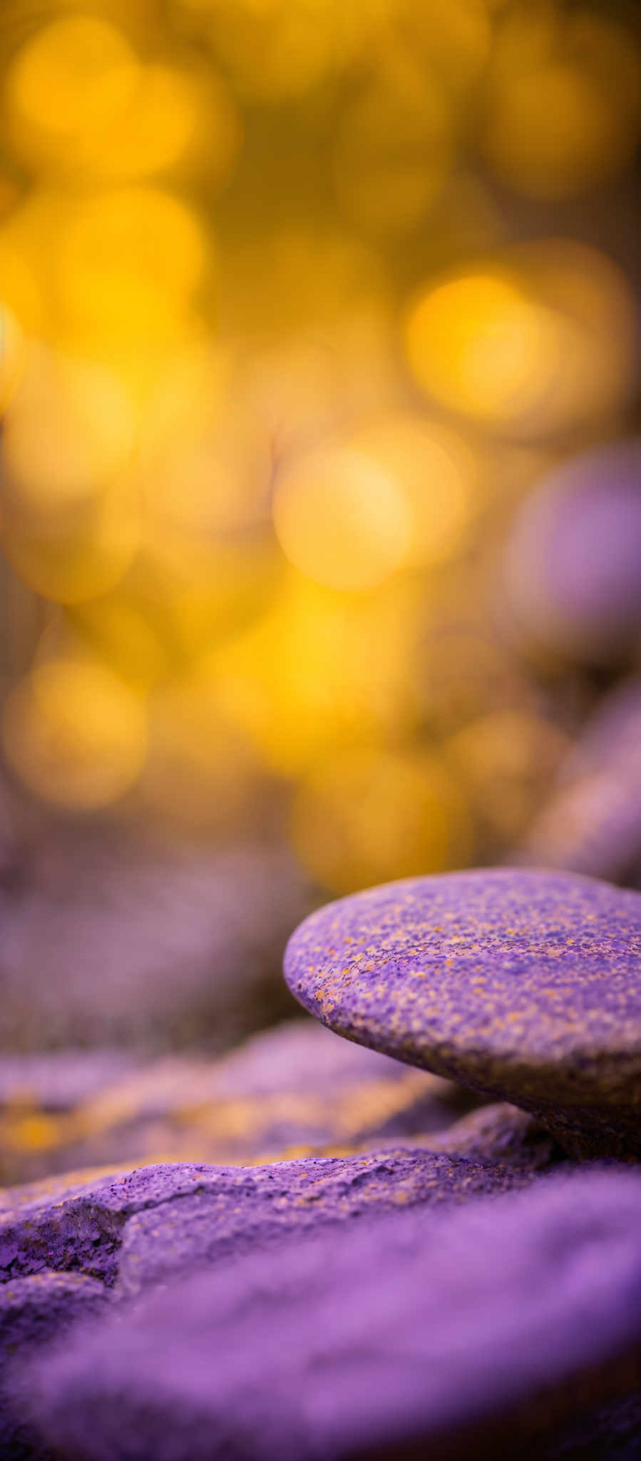 The image showcases a close-up view of smooth, rounded stones. The stones have a speckled texture, with a mix of purple and brown hues. The background is blurred with a golden-yellow bokeh effect, possibly representing sunlight or a light source. The overall ambiance of the image is serene and tranquil, evoking feelings of calmness and peace.