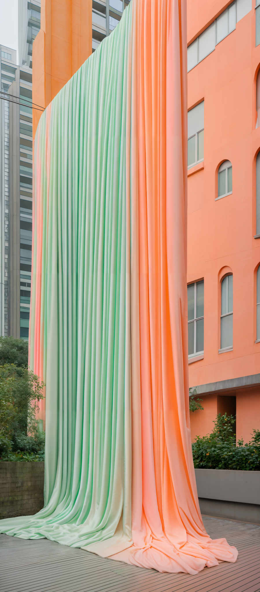 The image showcases a large, vertical installation of fabric that transitions in color from a light green at the top to a peachy-orange hue at the bottom. The fabric appears to be draped and cascades downwards, creating a soft, flowing effect. The background reveals a modern urban setting with buildings painted in various shades of orange and beige. There's also a hint of greenery, suggesting a courtyard or a garden area within the urban environment.