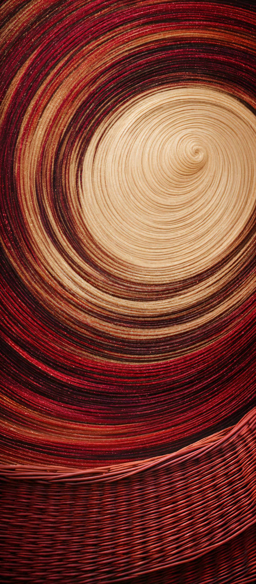 The image showcases a mesmerizing spiral pattern with a combination of vibrant colors. The predominant colors are shades of red, gold, and brown, creating a swirling effect. The spiral starts from a central point and expands outwards, with each layer exhibiting a unique texture and pattern. The bottom of the image displays a woven texture, possibly a basket or wicker, with a rich reddish-brown hue.