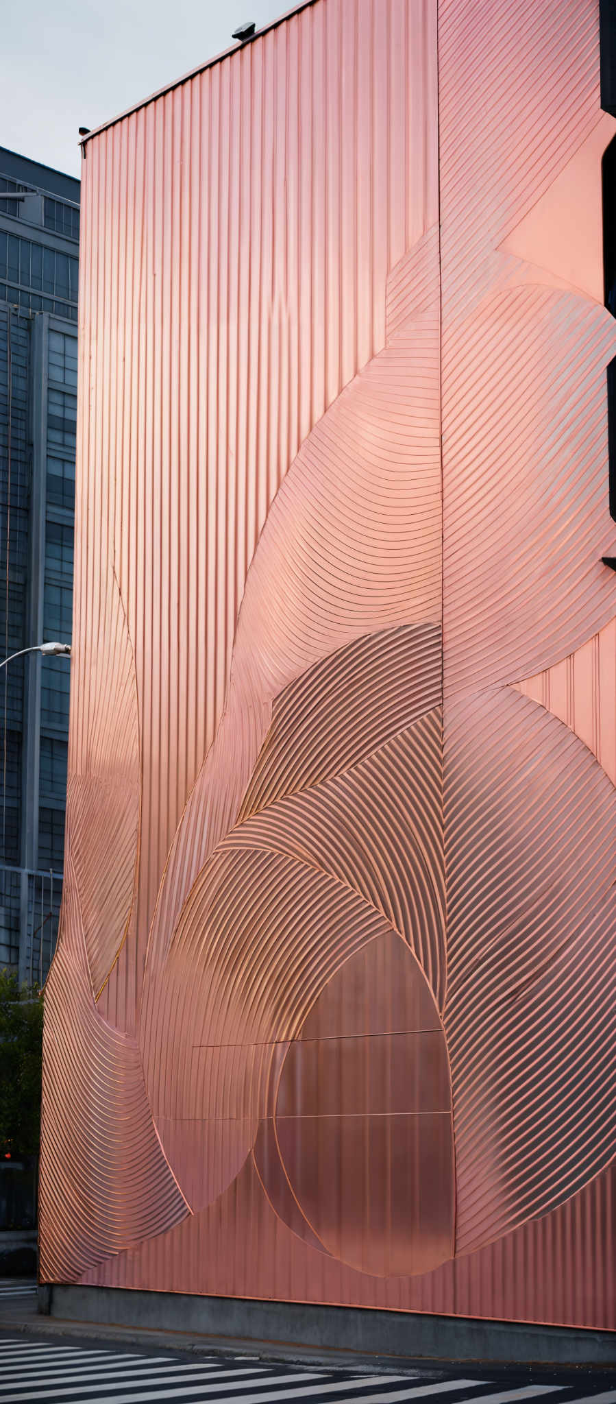 The image showcases a large building facade with a unique design. The dominant color is a shimmering rose gold or copper. The facade features intricate patterns of overlapping curves and lines, creating an abstract and artistic representation. The patterns give the appearance of waves or fluid motions. The building is adjacent to a street with a pedestrian crossing, and there are other buildings visible in the background.