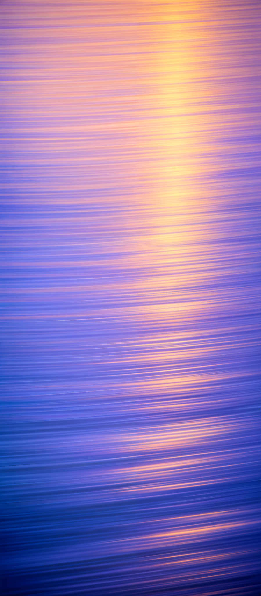 The image showcases a serene scene of a body of water, possibly a sea or ocean, during what appears to be either dawn or dusk. The water reflects a gradient of colors, transitioning from a deep blue at the top to a golden-orange hue near the horizon. The horizontal lines, possibly created by the movement of the water or a long exposure shot, add a rhythmic pattern to the image, creating a sense of calm and tranquility.