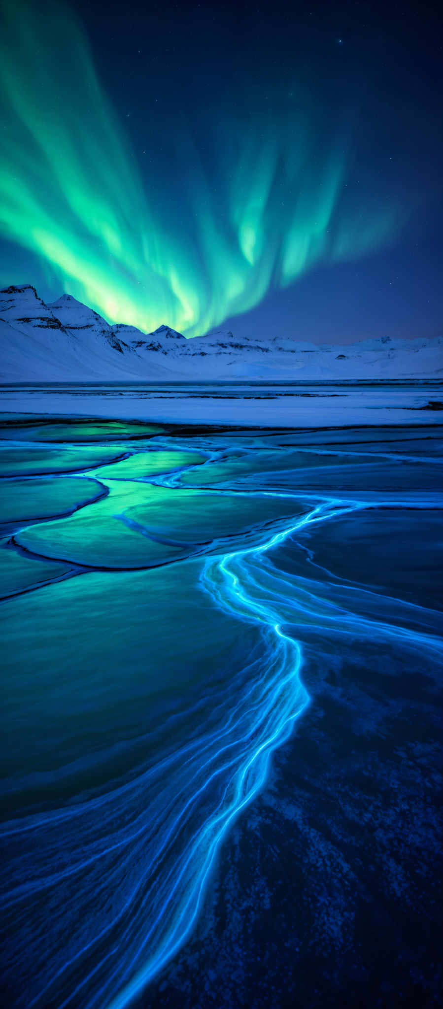 The image showcases a breathtaking view of the Northern Lights, also known as the Aurora Borealis. These lights display a vibrant shade of green, creating a mesmerizing pattern across the night sky. Below the sky, there's a snowy landscape with mountains in the distance. The foreground features a body of water with a unique pattern, possibly caused by the movement of the water or ice. The water reflects the green hues of the Auroras, creating an ethereal glow. The entire scene is illuminated by the natural light from the Auroral display, casting a serene and magical ambiance.