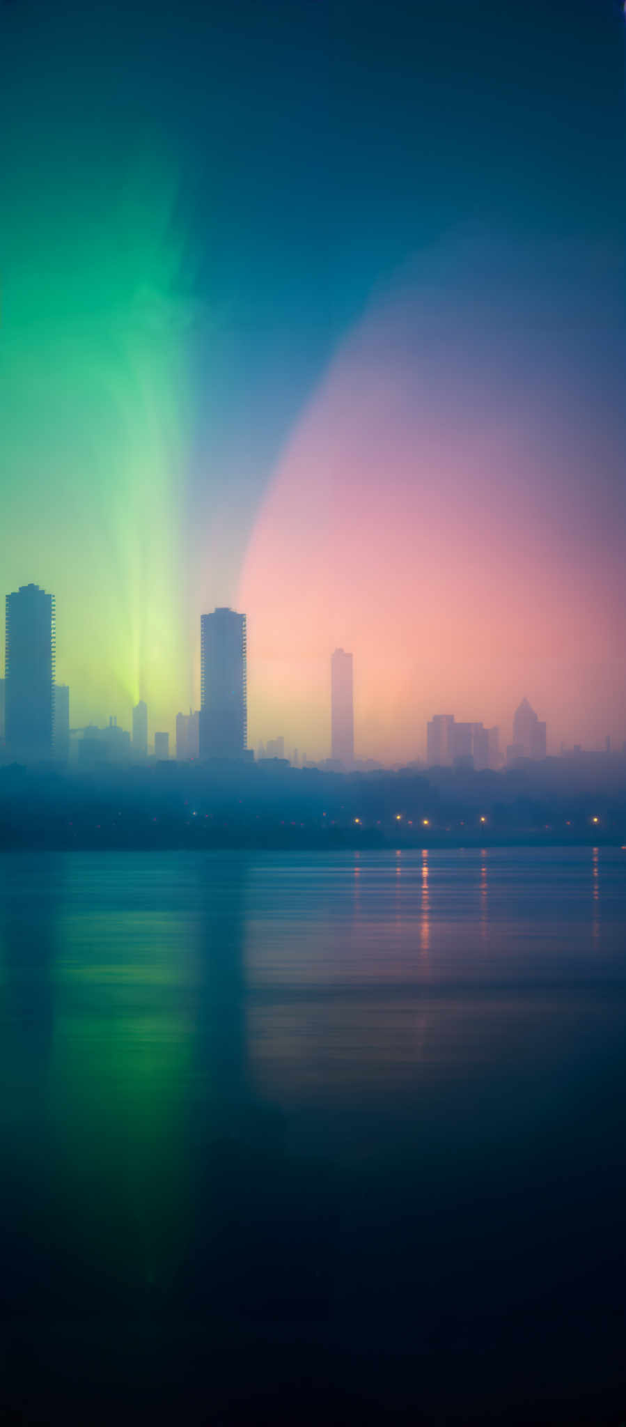 The image showcases a serene cityscape during what appears to be dawn or dusk. Dominating the foreground is a calm body of water reflecting the colors of the sky. The sky itself is a gradient of colors, transitioning from a deep blue at the top to a vibrant green at the bottom. This green hue is most likely caused by the sun's rays filtering through the atmosphere. The city skyline, visible in the middle, consists of tall buildings, some of which are illuminated from within. The buildings cast a soft glow on the water, creating a beautiful contrast with the ambient light. The overall mood of the image is tranquil and dreamy.