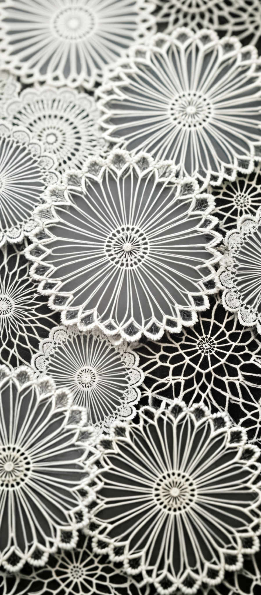 A close up of a white lace doily with intricate designs. The doily is composed of numerous small circles and lines creating a complex pattern. The design is symmetrical with the center of the doily being the most detailed. The edges of the circles are scalloped adding to the overall intricacy of the design. The color of the lace is pure white providing a stark contrast to the black background. The image is a close up allowing for a detailed view of the intricate design.