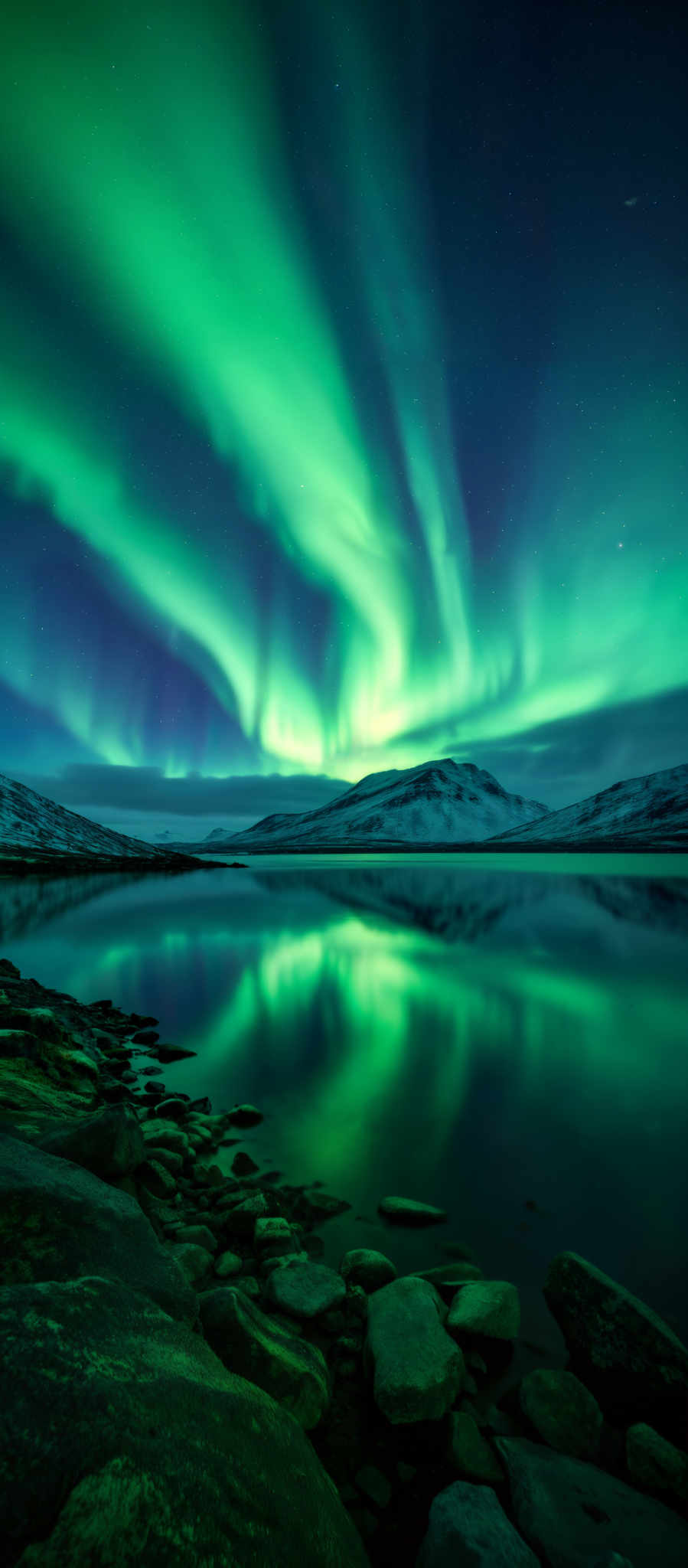 A serene scene of a lake with a mountain in the background. The lake is calm and still reflecting the bright green lights of the Aurora Borealis. The mountain covered in snow stands tall in the distance. The sky is dark providing a stark contrast to the vibrant colors of the natural phenomenon. The image captures the beauty and tranquility of nature with the Aurora lights adding a touch of magic to the scene.