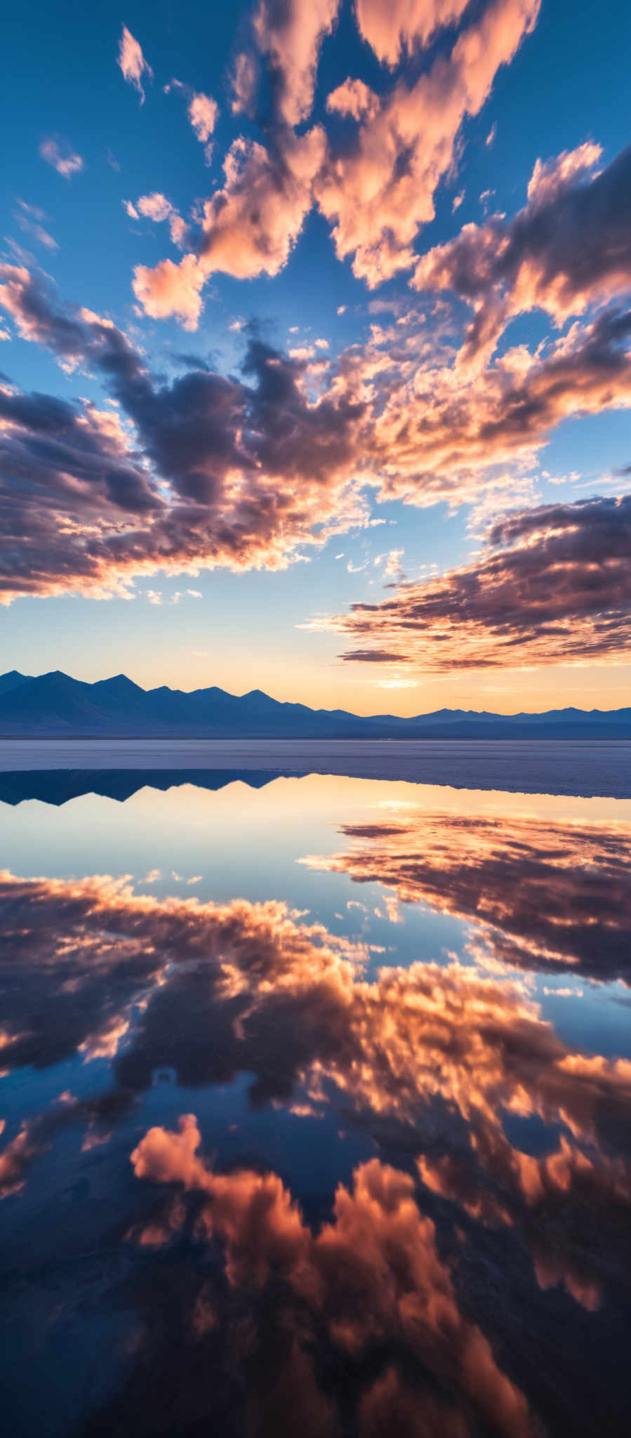 A serene landscape of a mountain range at sunset. The sky is painted with hues of orange and pink reflecting off the water below. The mountains appearing in the distance are bathed in a soft blue light. The water calm and still mirrors the sky and the mountains creating a beautiful symmetry. The image captures the tranquility and beauty of nature in its purest form.