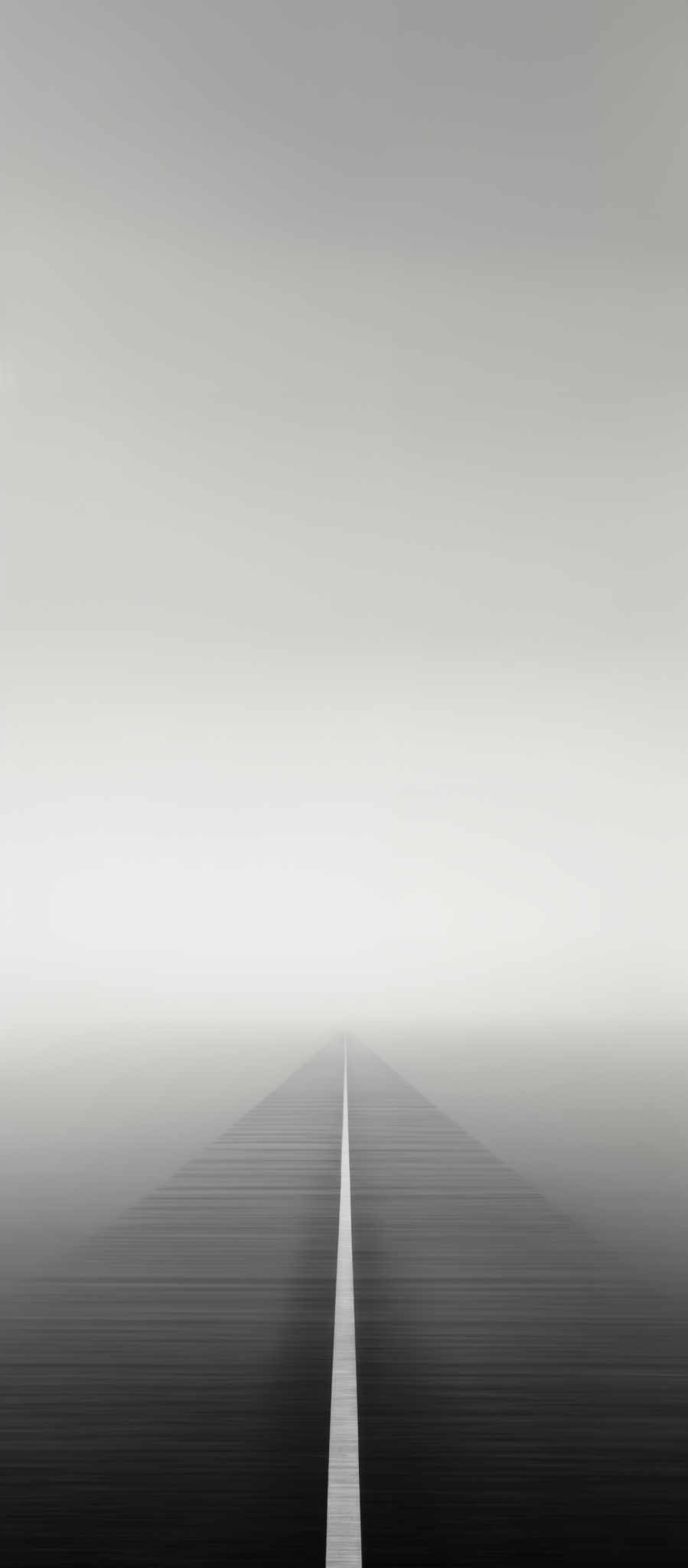 The image predominantly features shades of gray, creating a monochromatic appearance. The central focus is a straight, thin white line that extends from the bottom of the image towards the horizon. This line appears to be a pathway or a road leading into the distance. The surrounding area is filled with a soft, diffused fog or mist, making the horizon appear hazy and indistinct. The overall shape of the scene is elongated and symmetrical, with the pathway serving as a central vertical axis.