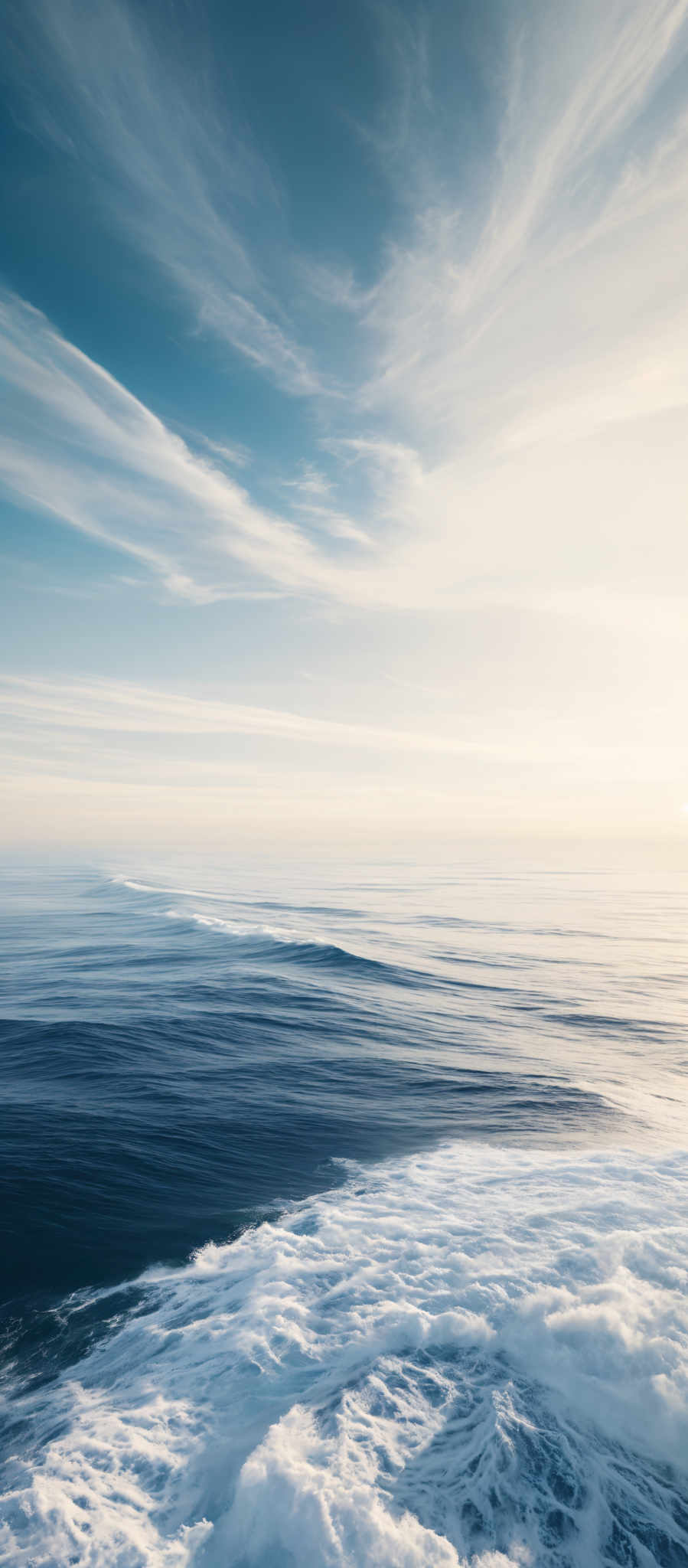 The image showcases a vast expanse of the ocean meeting the sky. The sky is painted with hues of blue and white, with wispy clouds stretching across. The ocean displays a deep blue color, transitioning to a lighter shade near the horizon. The waves are white, indicating their frothy nature, and they appear to be crashing near the bottom of the image, creating a foamy texture.