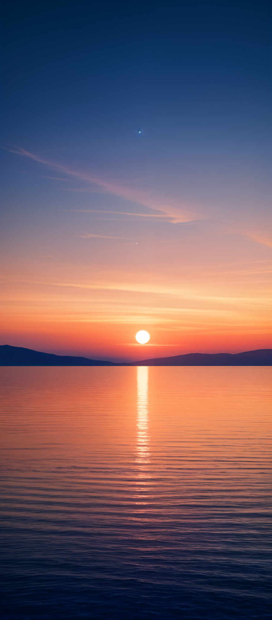 The image showcases a serene sunset over a calm body of water. The sky is painted with hues of deep blue transitioning to a vibrant orange and pink near the horizon. A few wispy clouds are scattered across the sky. The sun is captured just above the waterline, casting a golden reflection on the water's surface. The horizon reveals a faint silhouette of land or islands.