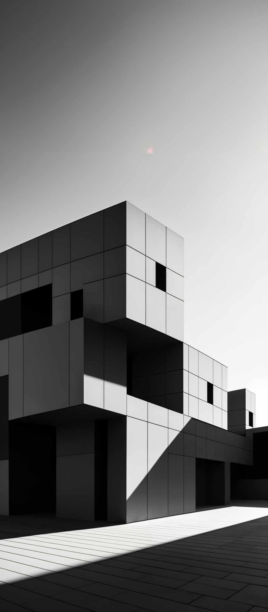 The image showcases a modern architectural structure with a predominantly monochromatic palette. The building is composed of interconnected cubes and rectangular blocks, each having varying sizes and orientations. The entire structure is enveloped in a sleek, reflective material, giving it a shiny appearance. The design is minimalistic, with clean lines and a geometric pattern. The image is in black and white, emphasizing the contrasts and shadows, which adds depth and drama to the scene.