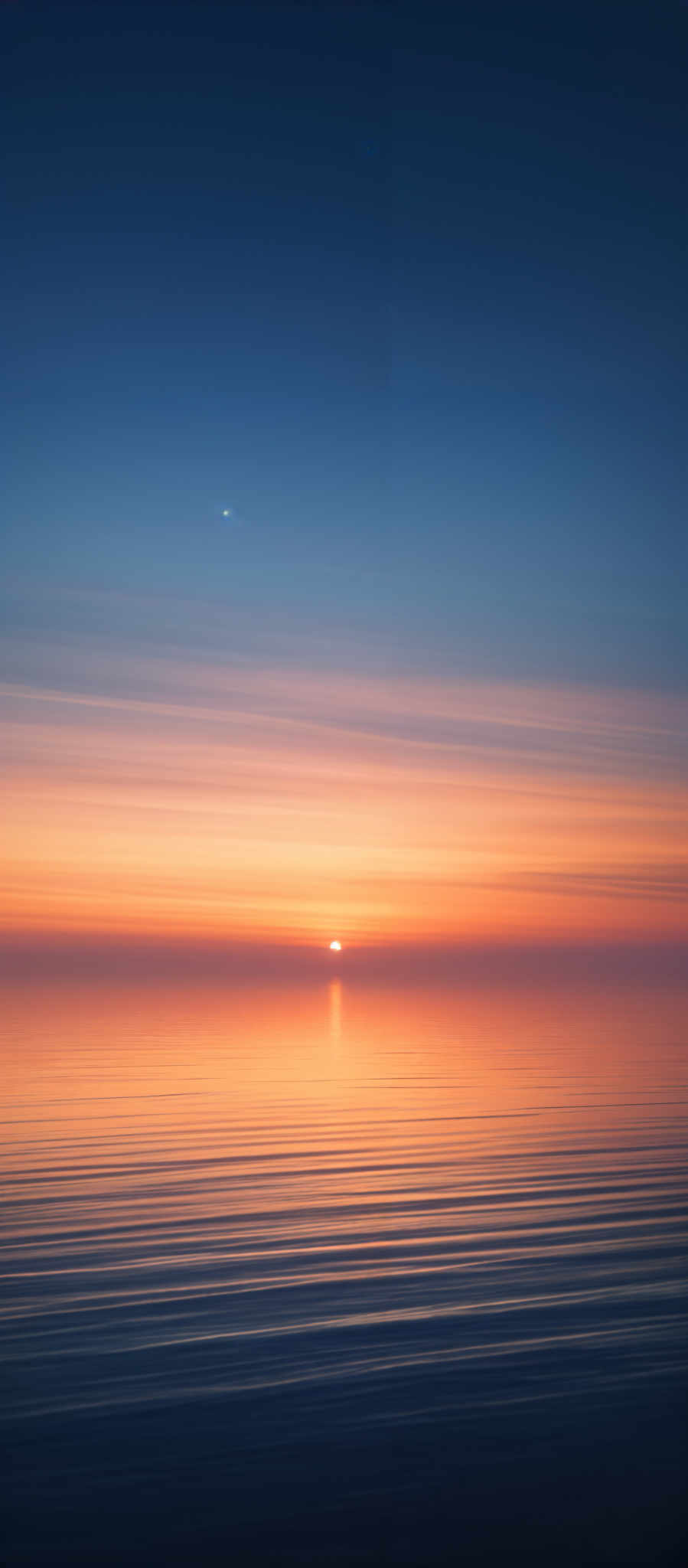 The image showcases a serene seascape during sunset. The sky transitions from a deep blue at the top to a gradient of orange and pink hues near the horizon. The sun is captured just above the waterline, casting a warm, golden glow. The water reflects the colors of the sky, creating a mirror-like effect. The horizon is defined by the meeting point of the sea and the sky. There are also subtle streaks in the sky that might suggest the movement of clouds or perhaps a long exposure effect.