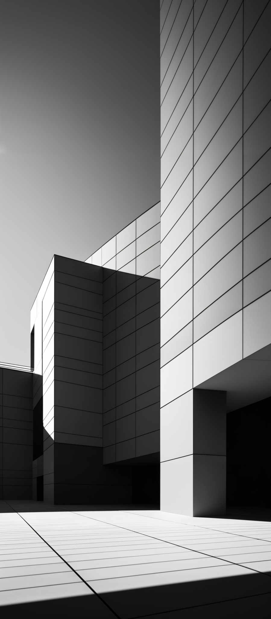 The image showcases a monochromatic architectural scene. The dominant colors are shades of gray, ranging from light to dark. The architectural design consists of geometric shapes, predominantly rectangles and squares, forming a modern building. The building's exterior is covered with a pattern of horizontal lines, possibly representing windows or panels. The image captures the building from a side angle, emphasizing its sharp angles and geometric forms. The shadows cast by the building' s structure create a contrast, adding depth and dimension to the photograph.