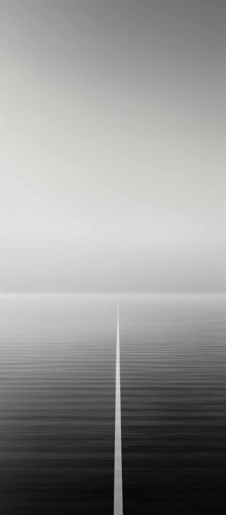 The image is in black and white, showcasing a vast expanse of calm water with a straight, thin white line running vertically down the center. The horizon is clearly visible, dividing the water from the sky. The sky is vast and appears to be cloudless, with a gradient of lightness from the top to the bottom. The water reflects the sky's light, creating a serene and symmetrical visual effect.