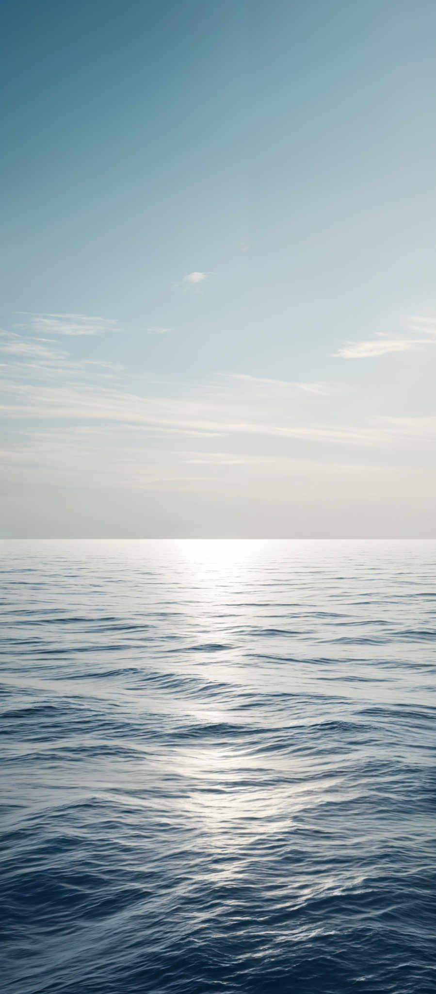 The image showcases a vast expanse of calm sea with gentle ripples. The sky above is clear with a gradient of light blue, transitioning to a deeper shade near the horizon. The sun is visible, casting a radiant glow over the water, creating a shimmering reflection. The horizon line is distinct, separating the serene sky from the tranquil sea.