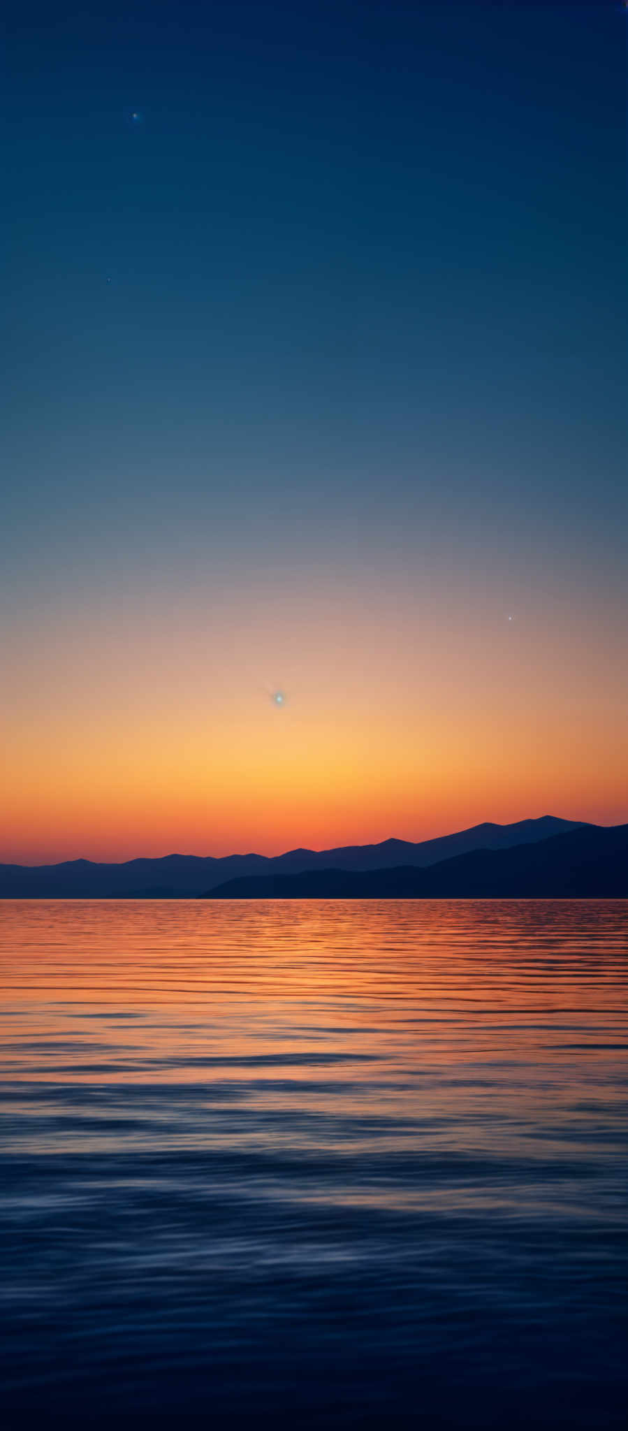 The image showcases a serene landscape during what appears to be either dawn or dusk. The sky transitions from a deep blue at the top to a warm orange near the horizon. There are three celestial bodies visible: two planets or moons and a bright star or planet. The horizon is marked by a silhouette of a mountain range. The water below reflects the colors of the sky, creating a gradient of blues and oranges. The calm water surface has gentle ripples, suggesting a gentle breeze.