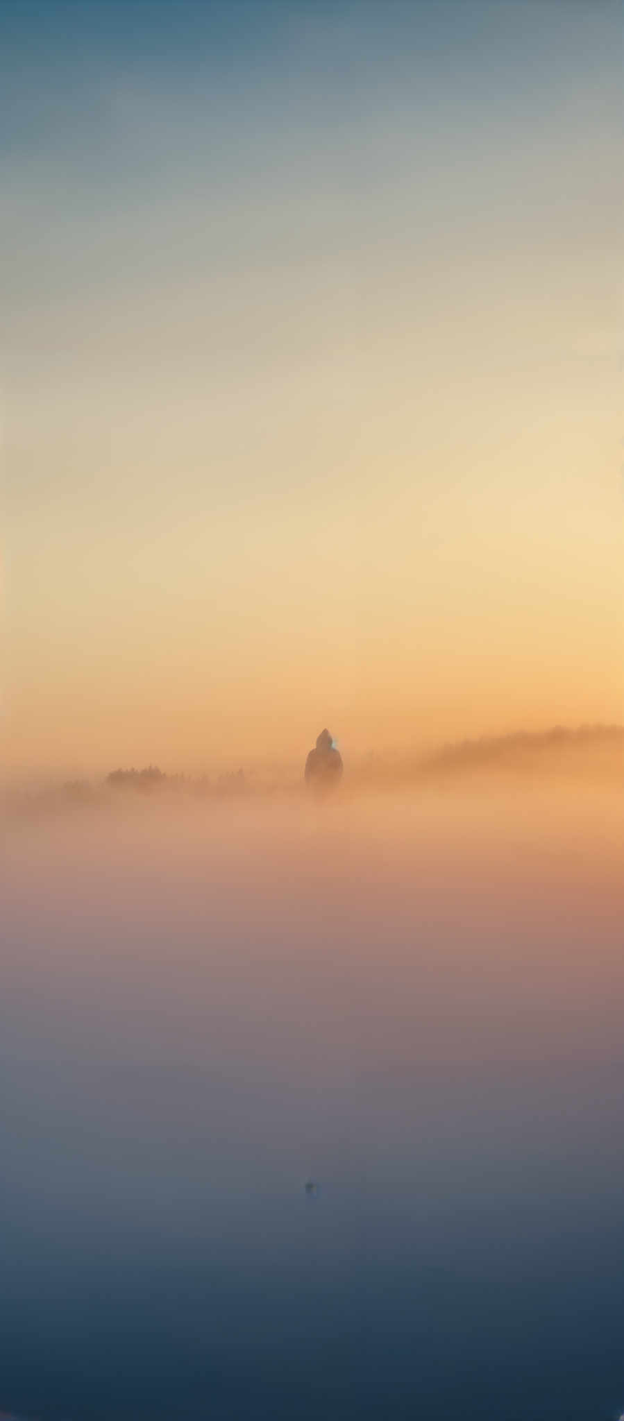 The image showcases a serene landscape during what appears to be either dawn or dusk. The sky is painted with hues of orange, yellow, and blue, transitioning smoothly from one to the other. Below the sky, a thick layer of mist or fog blankets the ground, creating a dreamy atmosphere. In the distance, a lone structure, possibly a building or a tower, stands amidst the fog, its silhouette dark against the luminous sky. The structure has a dome-like top, suggesting it might be a church or a similar edifice. The water below reflects the colors of the sky and the mist, adding to the tranquility of the scene.