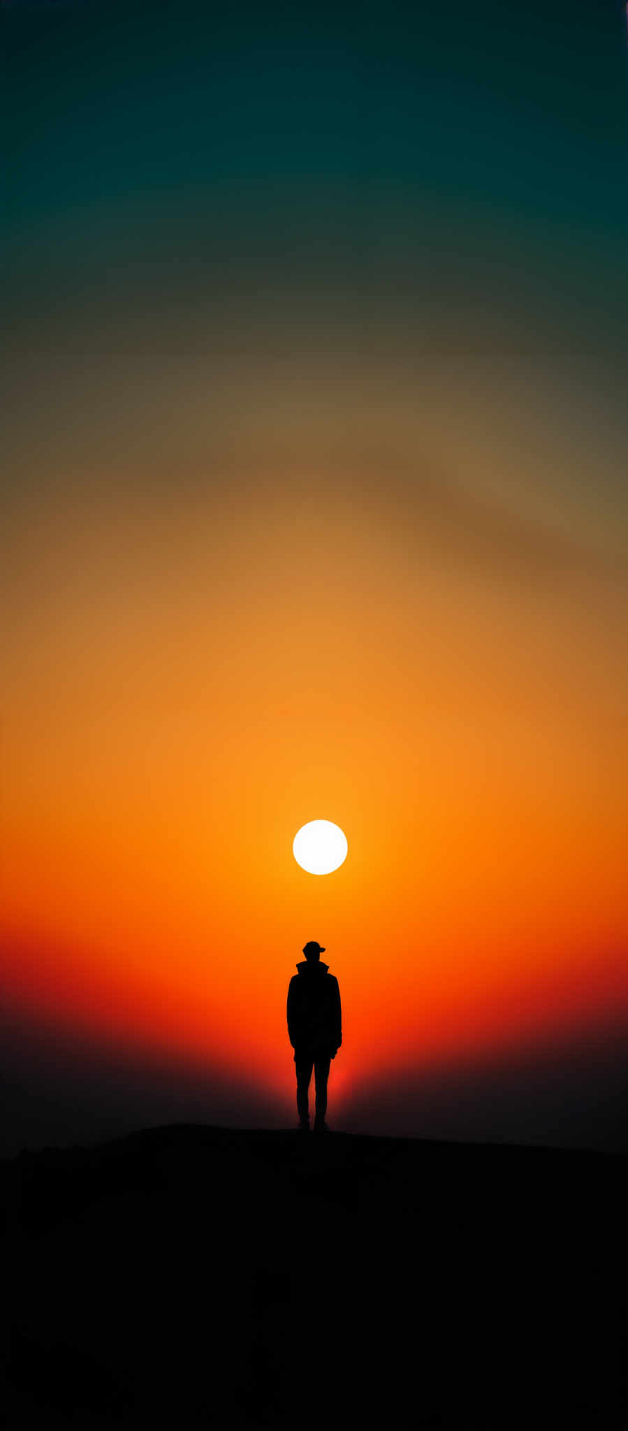 The image showcases a breathtaking sunset with a gradient of colors ranging from deep blue at the top to fiery orange and red at the bottom. The sun is positioned centrally, casting a bright glow and creating a silhouette effect on the horizon. In the foreground, there's a silhette of a person wearing a hat, standing on what appears to be a hill or elevated ground. The person seems to be gazing at the sunset, adding a sense of contemplation and solitude to the scene.