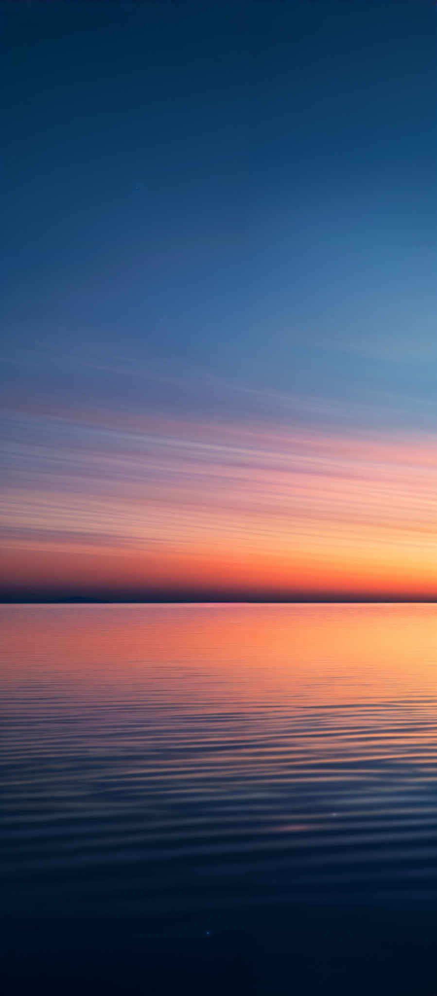 The image showcases a serene seascape during what appears to be either dawn or dusk. The sky is painted with hues of deep blue transitioning to lighter shades, and a few streaks of white, possibly representing clouds or stars. The horizon reveals a vibrant gradient of colors, ranging from deep purples and blues to fiery oranges and yellows. Below the horizon, the calm sea reflects these colors, creating a mirror-like effect. The gentle waves on the water's surface add texture and depth to the scene.