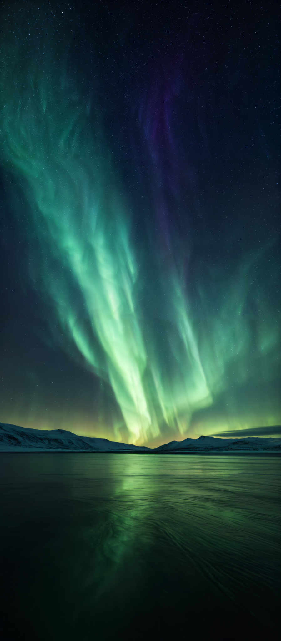 The image showcases a breathtaking view of the Northern Lights, also known as the Aurora Borealis. The colors range from vibrant green to deep purple, creating an ethereal dance across the night sky. The shape is predominantly curvilinear, with the lights flowing and swirling in a manner reminiscent of waves or flames. The celestial display is reflected in a calm body of water below, creating a mirror effect. The horizon is dotted with snow-covered landforms, and the vast expanse of the starry sky adds to the overall majesty of the scene.