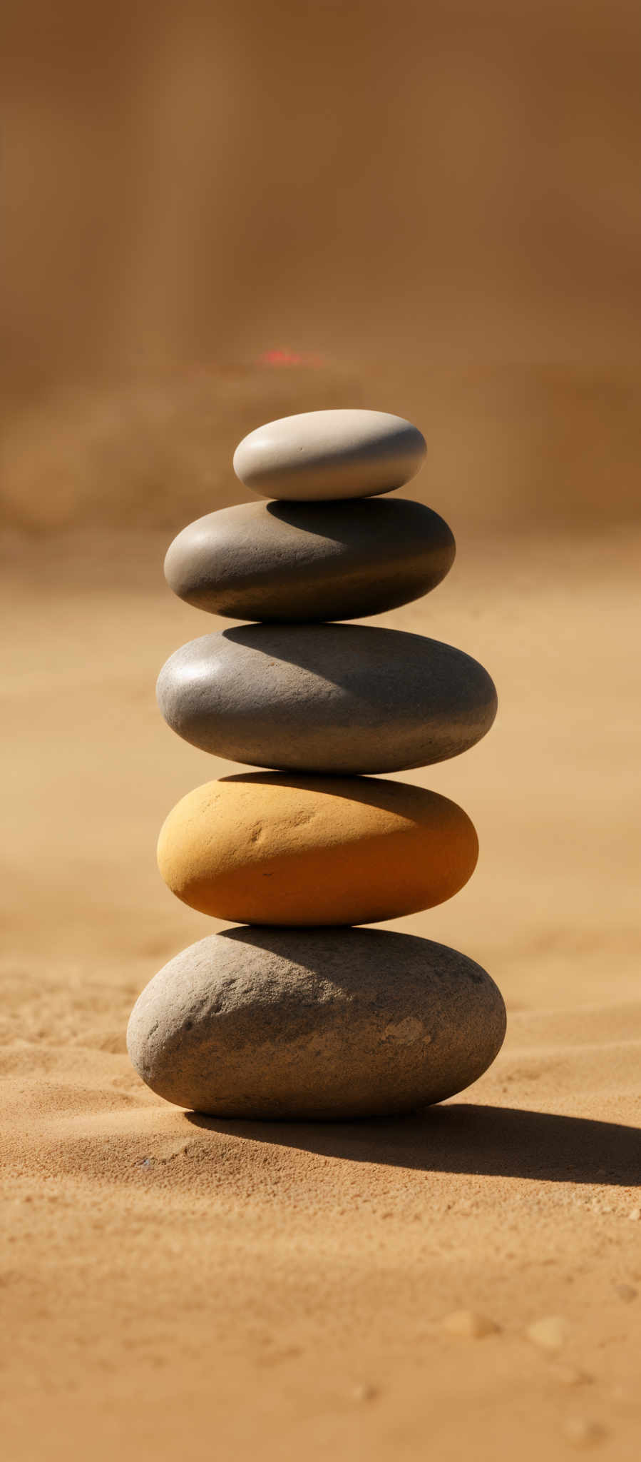 The image showcases a stack of smooth, rounded stones set against a sandy background. The stones vary in color, with shades of gray, beige, and a hint of orange. The stack is balanced, with each stone resting on the one below it, creating a harmonious and serene visual.