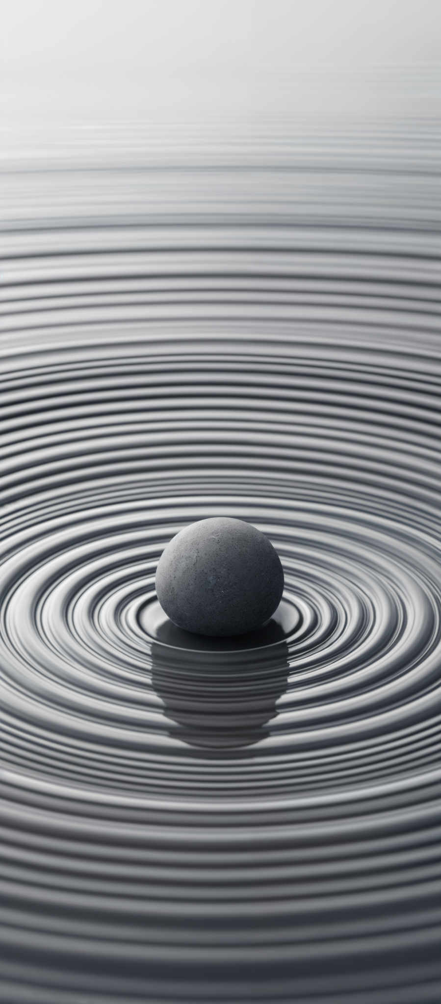 The image showcases a monochromatic palette with varying shades of gray. The primary focus is a smooth, oval-shaped stone placed in the center of concentric ripples in water. The ripple patterns radiate outward from the stone, creating a serene and symmetrical effect. The stone appears to be of a muted gray color, and the water reflects the stone's hue, adding depth to the scene.