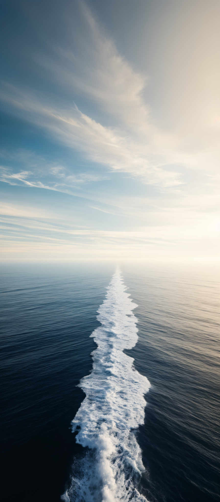 The image showcases a vast expanse of the ocean with a wake trail of a ship or boat. The wake is a white, frothy path that extends from the ship's rear, cutting through the calm blue waters. Above, the sky is painted with hues of blue and white, with wispy clouds stretching across. The horizon is faintly visible, and the overall mood of the image is serene and tranquil.