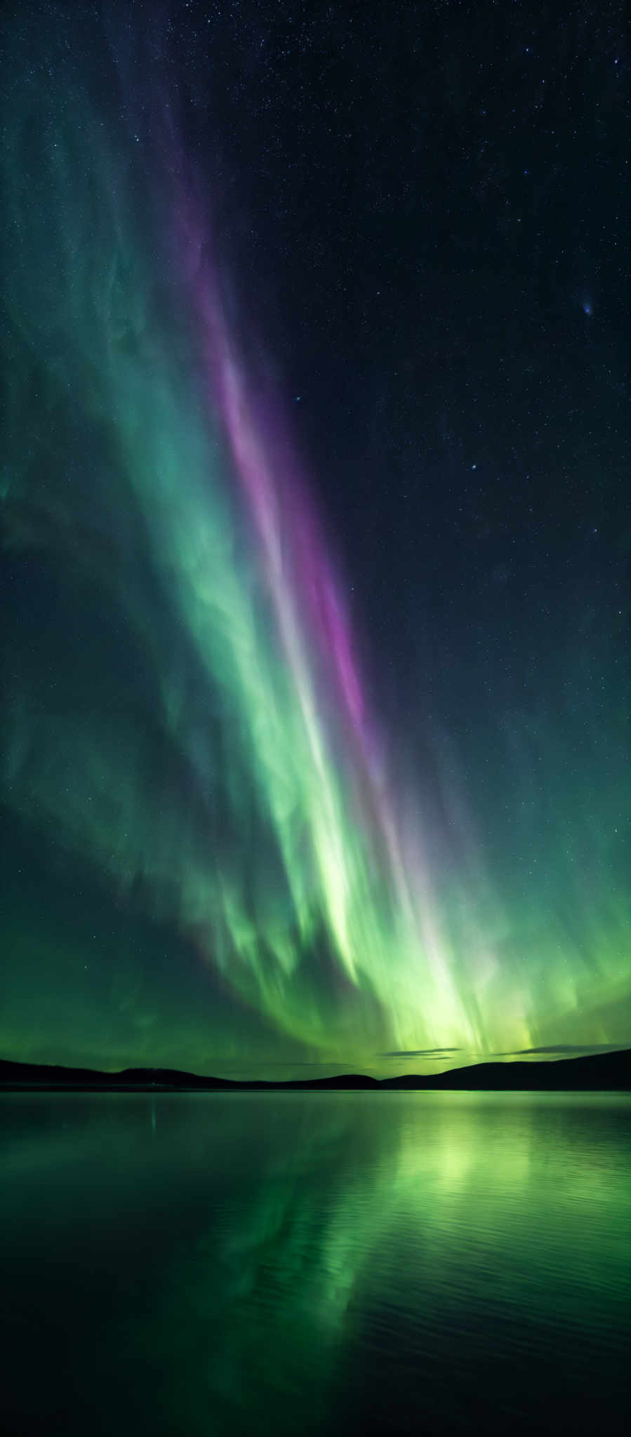 The image showcases a breathtaking view of the Northern Lights, also known as the Aurora Borealis. These lights display a vibrant array of colors, including deep blues, purples, and bright greens. The shape of the lights is wavy and seems to emanate from a central point, cascading outwards in both directions. The lights are reflected beautifully on a calm body of water below, creating a mirror-like effect. The sky is dotted with numerous stars, adding to the ethereal beauty of the scene.