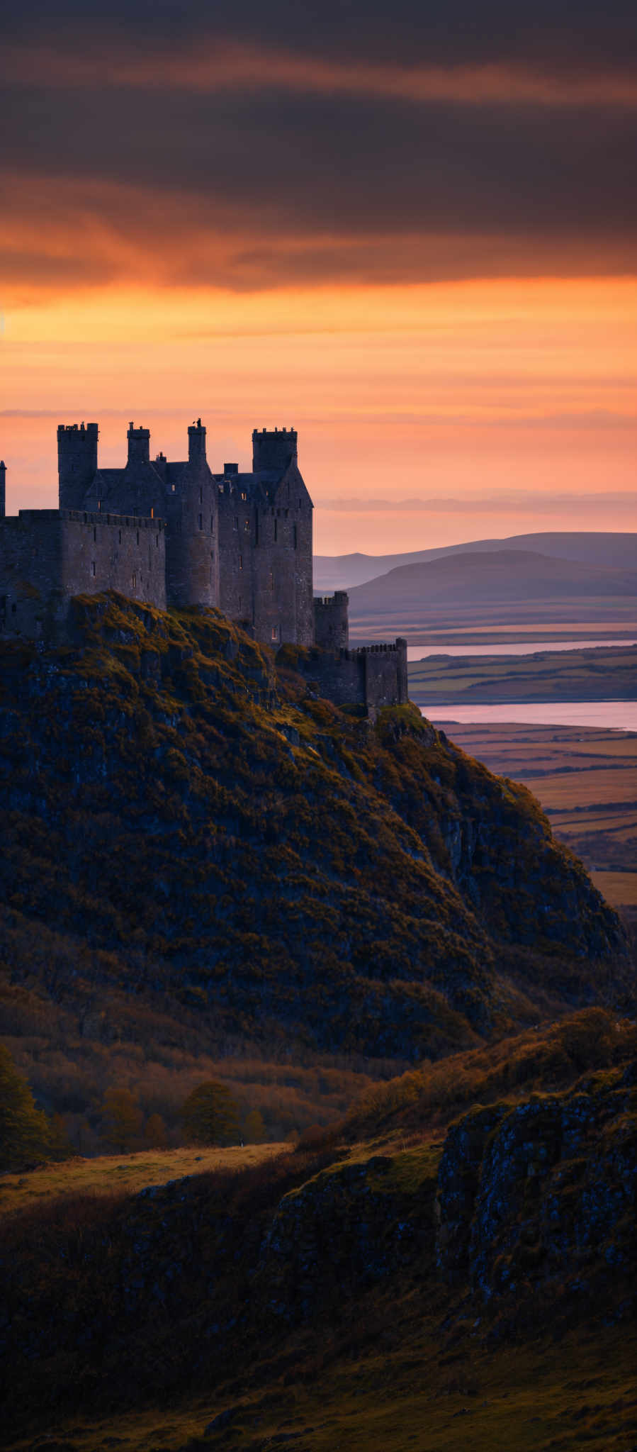 The image showcases a majestic castle perched atop a rugged hill. The castle is rectangular in shape with multiple turrets and battlements. The hill is covered in greenery, with patches of moss and grass. The sky above is dramatic, painted in hues of orange, yellow, and gray, suggesting either a sunrise or sunset. In the distance, there are flatlands with a river or a large body of water reflecting the sky's colors.