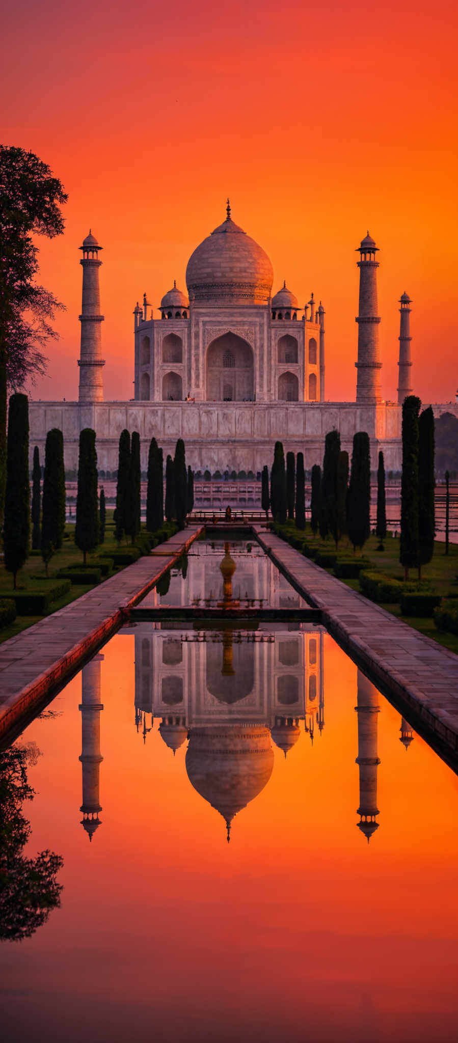 The image showcases a breathtaking view of the Taj Mahal during sunset. The sky is painted in vibrant shades of orange and red, creating a dramatic backdrop for the white marble mausoleum. The Taj is symmetrically flanked by two minarets on either side. In the foreground, there's a serene reflecting pool that mirrors the Tah Mahal and the surrounding gardens. The gardens are meticulously manicured with neatly trimmed trees and shrubs. The symmetry of the scene is further emphasized by the perfectly aligned pathways leading up to the Taja Mahal.