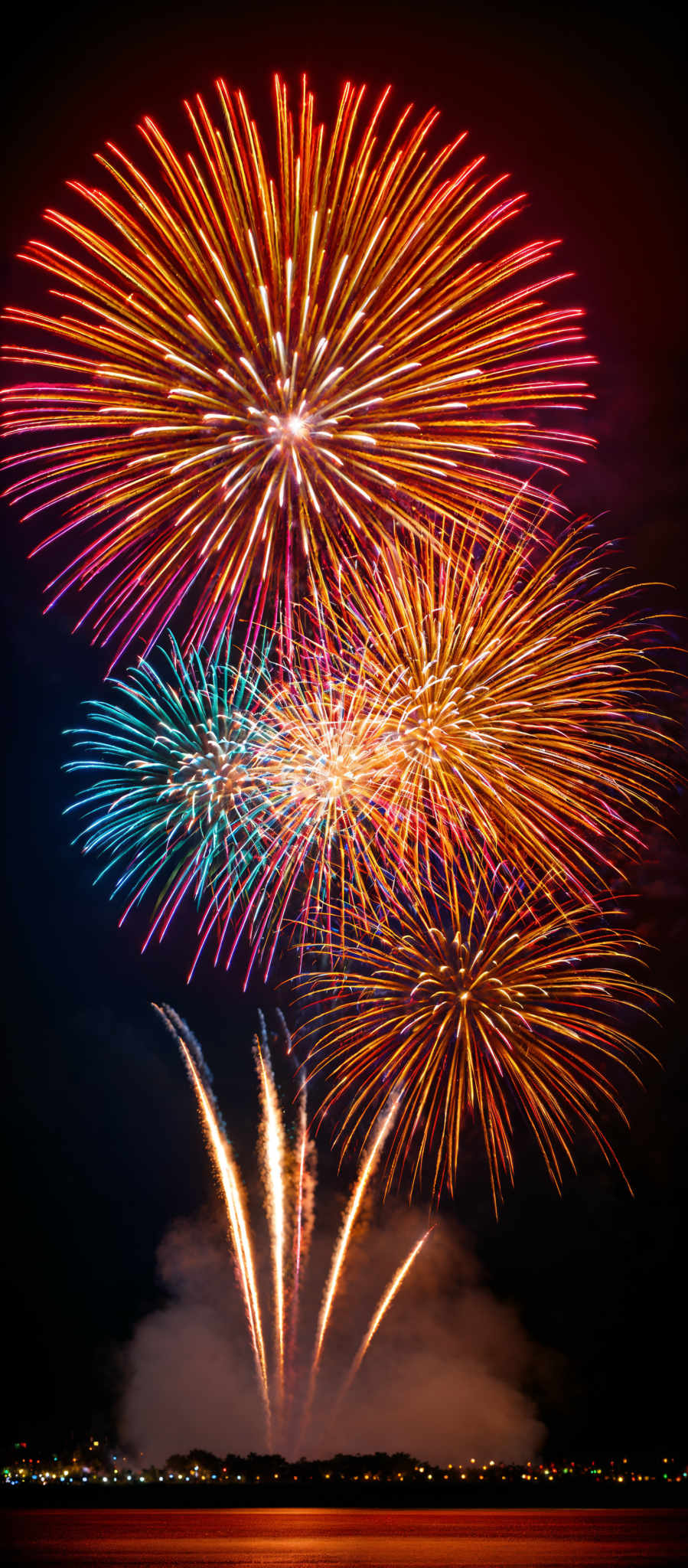 The image showcases a vibrant display of fireworks. The fireworks burst in a myriad of colors including red, blue, yellow, and pink. The shapes of the fireworks are predominantly spherical with some exhibiting starburst patterns. The bursts are spread out, creating a radiant effect against the dark backdrop of the night sky.