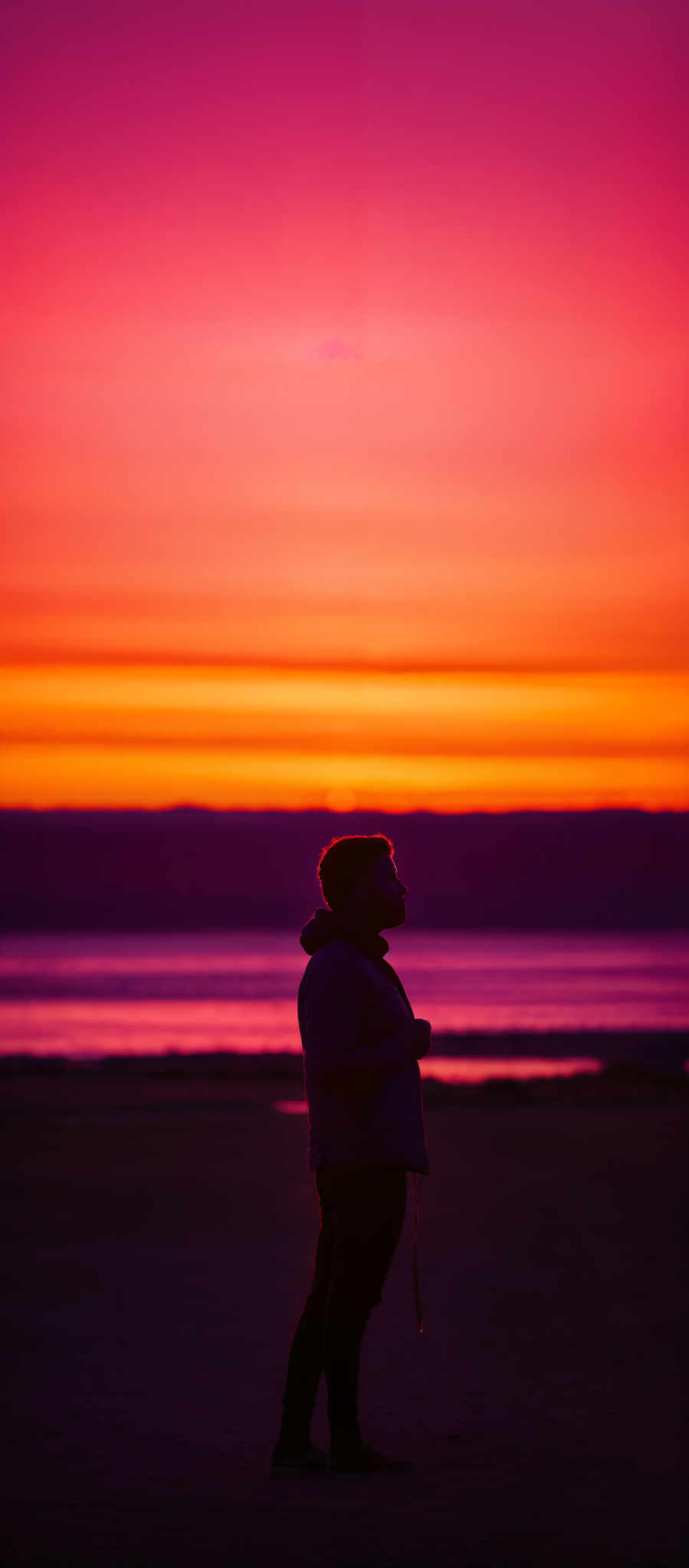 The image showcases a vibrant sunset with hues of pink, orange, and purple. The sky transitions from a deep pink at the top to a fiery orange near the horizon. Below the horizon, there's a hint of a darker shade, possibly indicating the onset of night. In the foreground, there is a silhouette of a person standing on what appears to be a beach. The person is facing the sunset, and their profile is illuminated by the warm glow. The individual is wearing a jacket and appears to have a contemplative expression. The beach stretches out to the right, with the calm sea reflecting the colors of the sky.