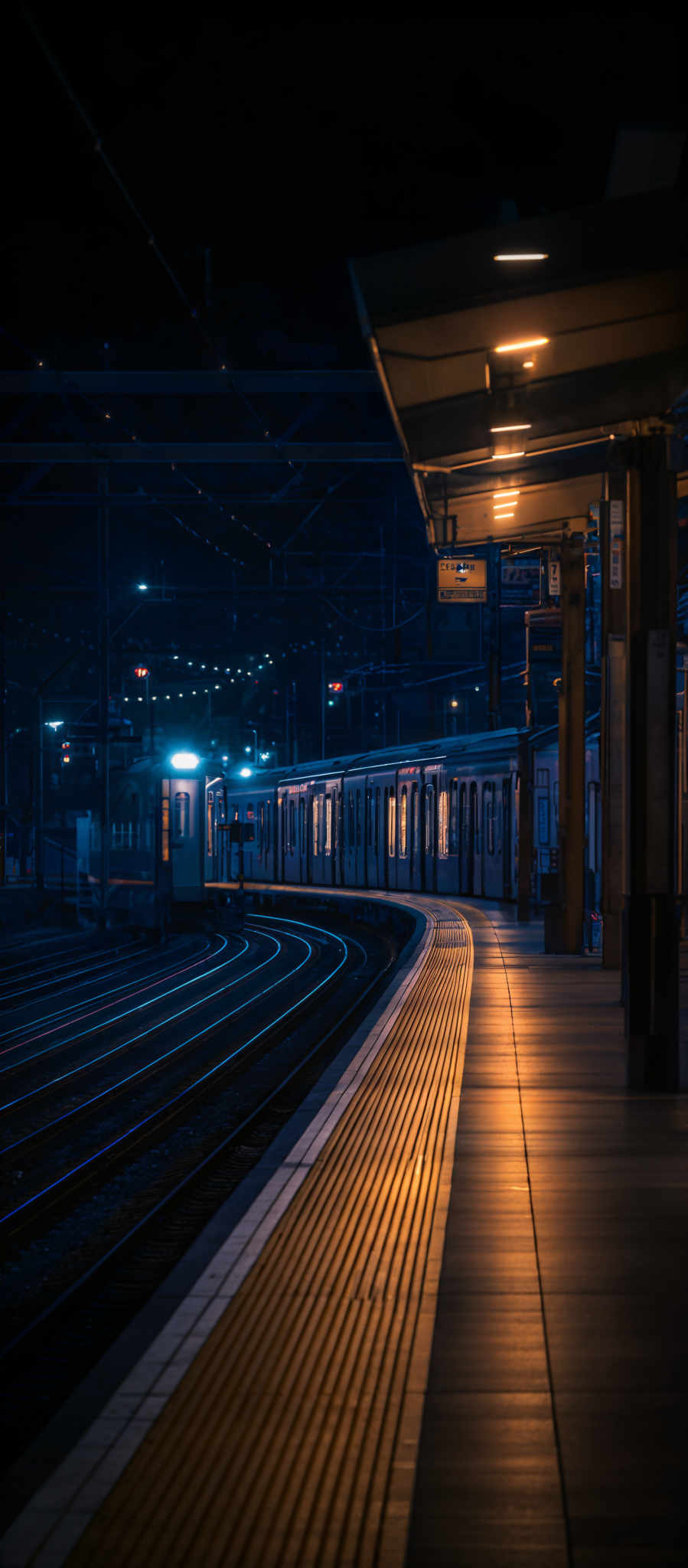 The image showcases a nighttime scene at a train station. The dominant colors are deep blues and warm yellows. The train station platform is curved, with a series of yellow tactile paving strips running along its length. Above the platform, there are overhead lights illuminating the area. The tracks are curved and parallel, with multiple train cars visible. The station has a modern design, with sleek structures and signage.