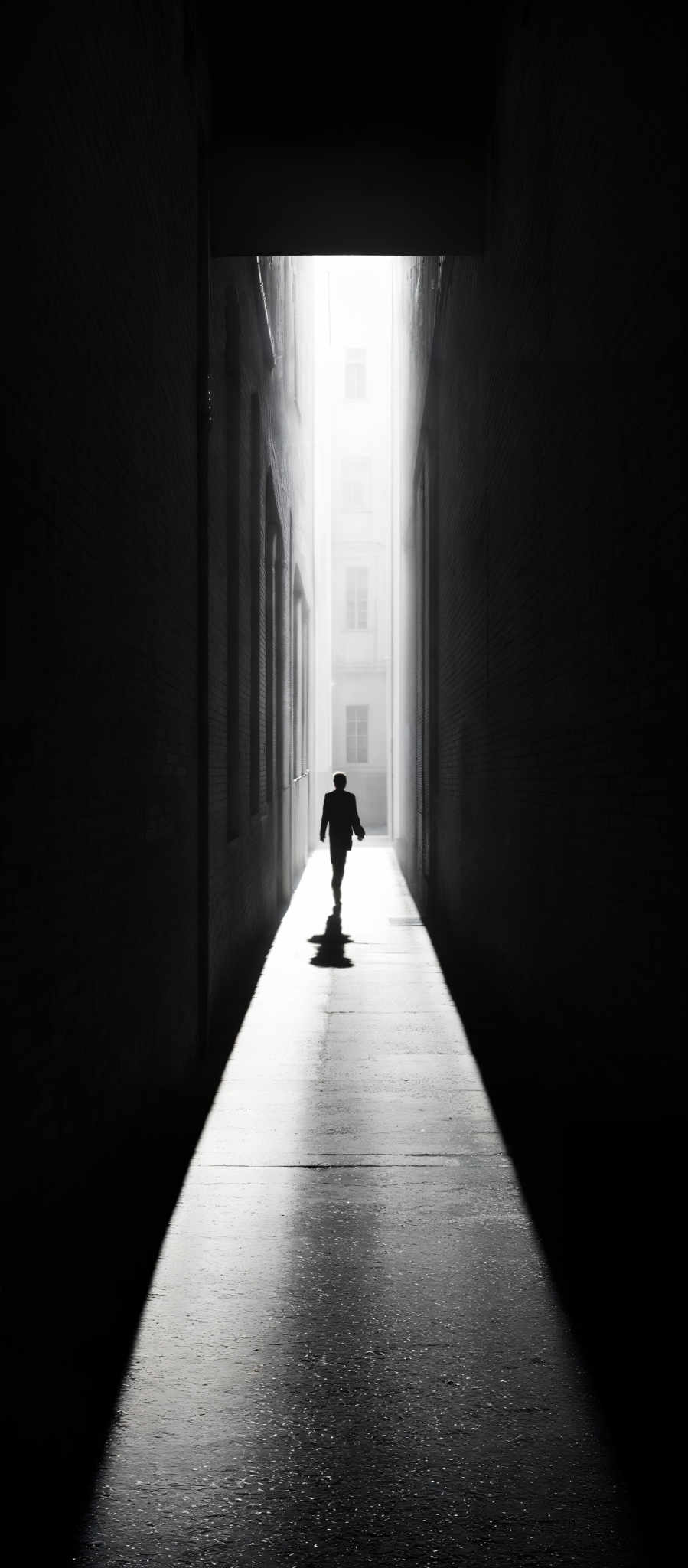 The image is in black and white, showcasing a narrow alleyway between tall buildings. The walls of the buildings are made of brick, and they are tall and narrow, creating a confined space. The alley is illuminated by a light source from the end, casting a long shadow of a person walking towards the light. The person appears to be wearing a hat and coat, suggesting a colder climate. The ground is smooth, possibly made of concrete, and the light creates a stark contrast between the dark walls and the bright pathway.