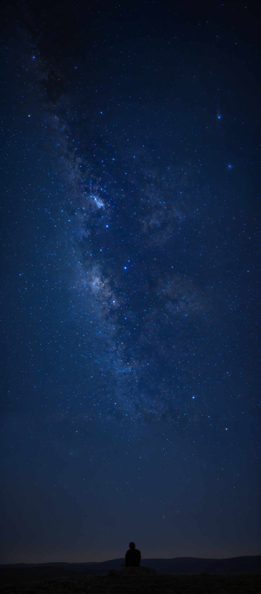 The image showcases a vast expanse of the night sky, dominated by a brilliant display of stars and the Milky Way galaxy. The Milky way appears as a dense, cloudy band of light, with countless stars shining brightly. The colors range from deep blues to lighter whites, with some areas appearing darker due to the dense concentration of stars. On the bottom right, there's a silhouette of a person sitting on a rock, seemingly gazing at the celestial spectacle, providing a sense of scale to the vastness of the universe.