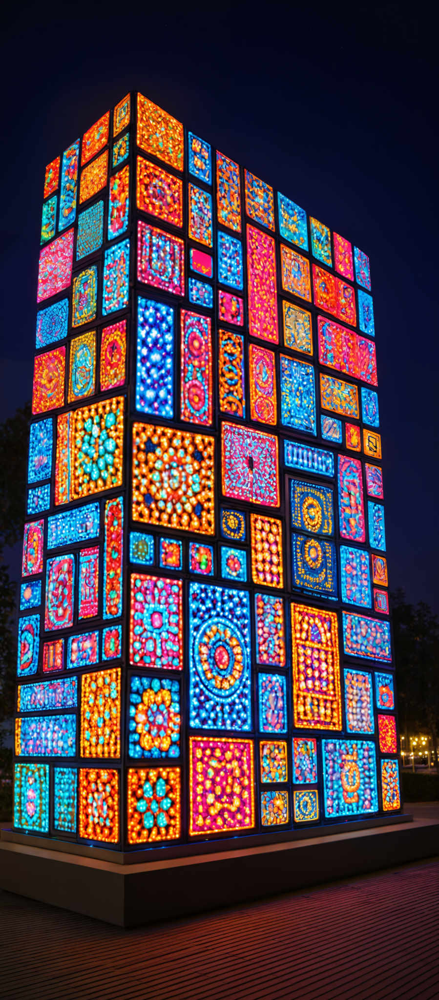 The image showcases a large structure or installation made up of numerous smaller panels. Each panel displays a vibrant and intricate design, predominantly featuring patterns and motifs in hues of blue, red, orange, and yellow. The designs are diverse, ranging from floral patterns to geometric shapes, and they appear to be illuminated, giving the entire structure a luminous and radiant appearance.