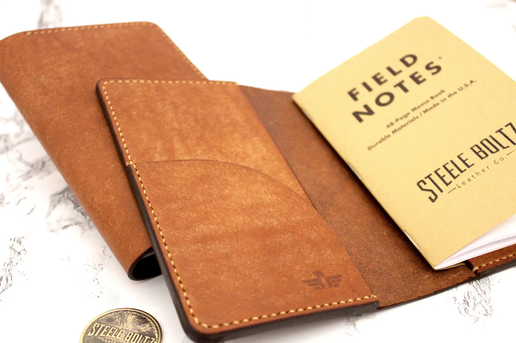 Leather notebook cover with "Field Notes" memo book and Steele Bolt logo coin on marble.
