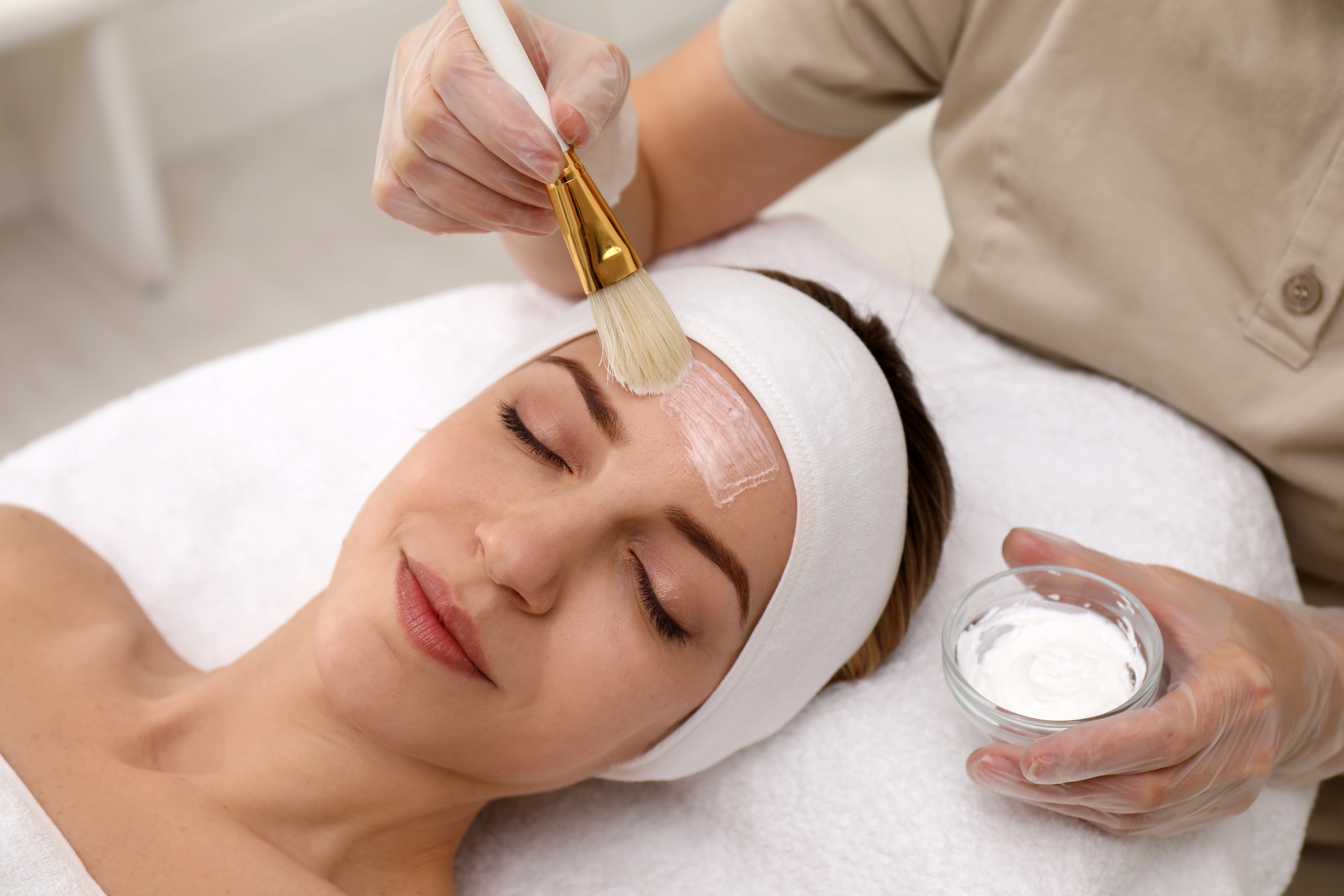 An esthetician applies a chemical peel solution to a relaxed woman's forehead using a brush during a facial treatment