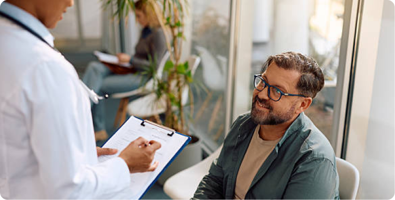 A doctor holding a clipboard consults with a smiling male patient in a bright waiting area.