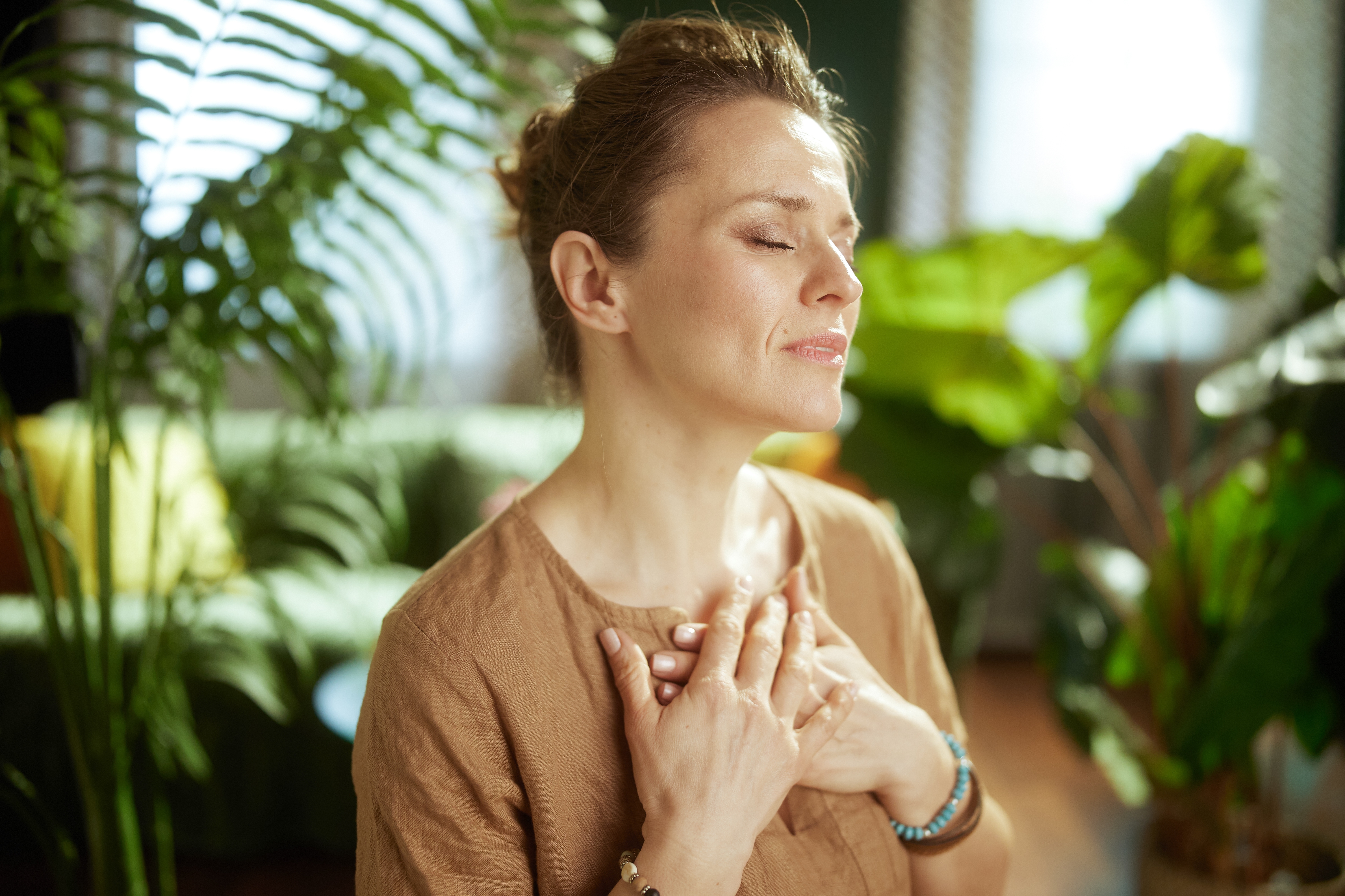 A woman stands with her eyes closed and hands clasped over her heart in a plant-filled living room, practicing calm breathing